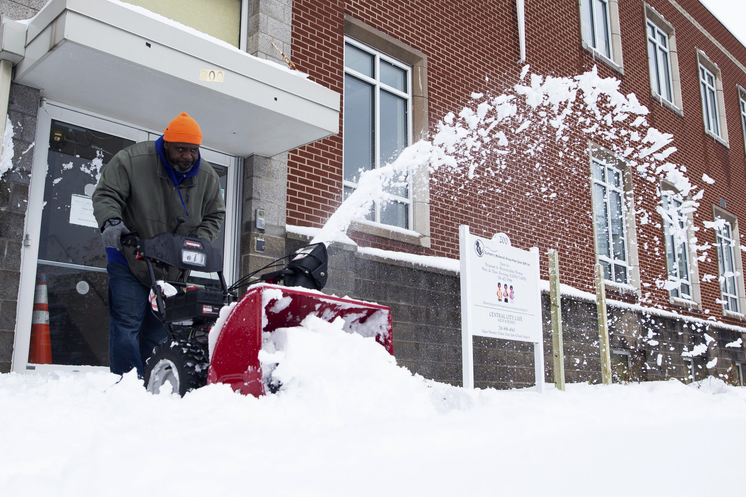 A man uses a snowblower to clear the sidewalk. There's a considerable amount of snow on the ground.