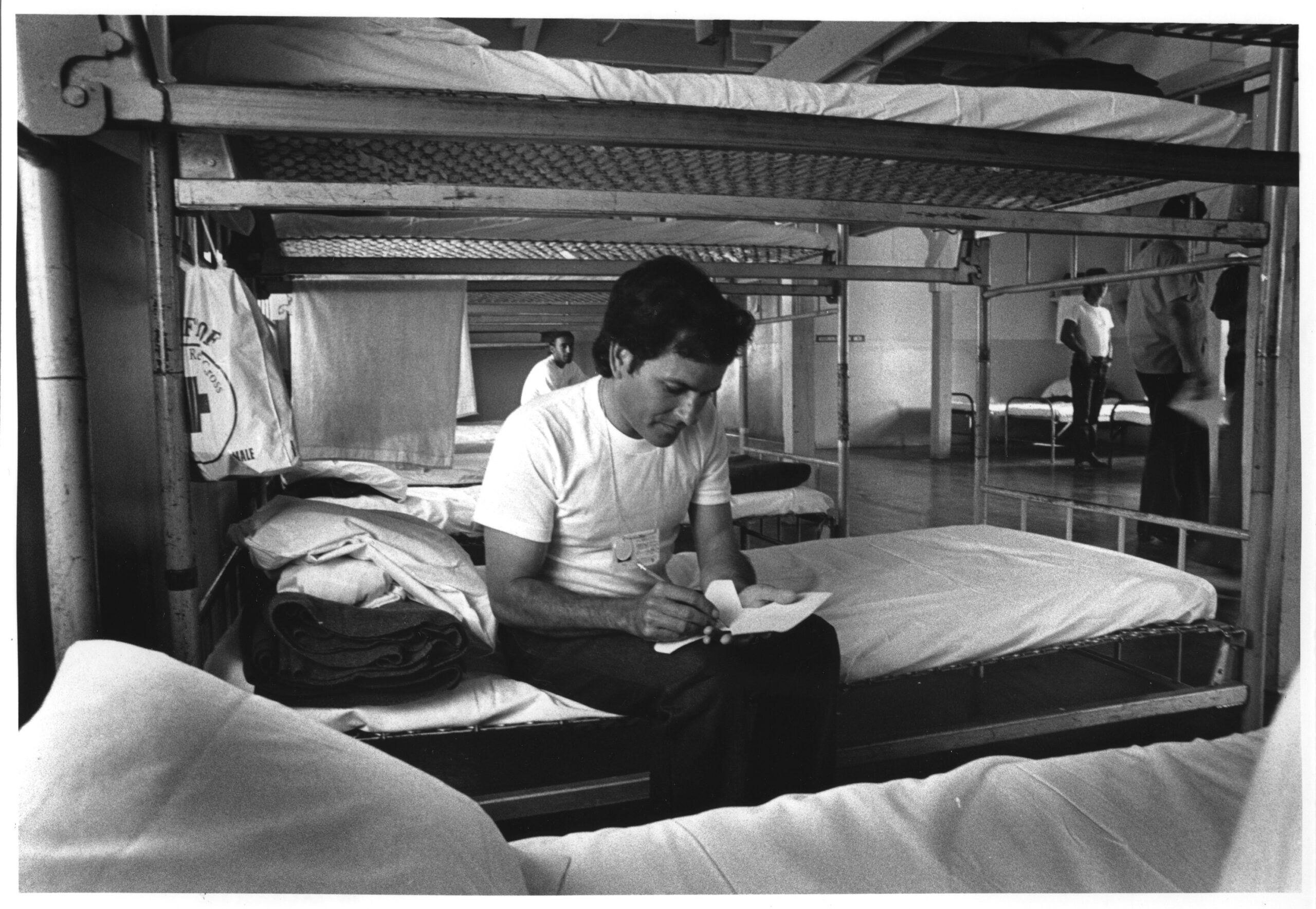 A black and white photograph of Carlos Miguel Larrosa Perez writing while sitting on a bed inside one of the barracks