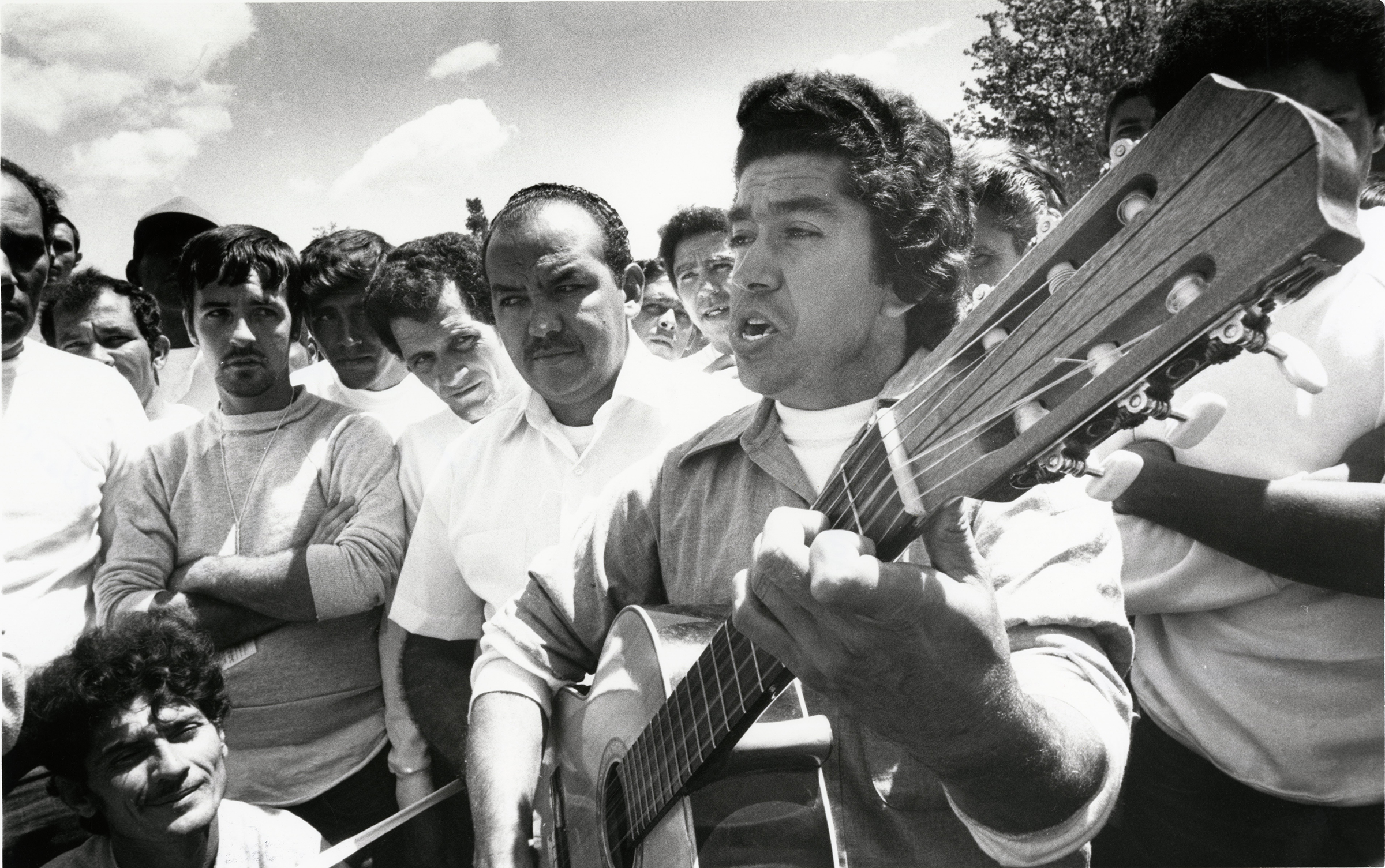 A black and white photograph of Ramiro Matos Nao playing guitar for a group of refugees