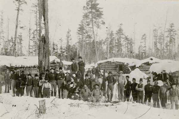 Logging crew posed in front of buildings at a logging camp near Antigo