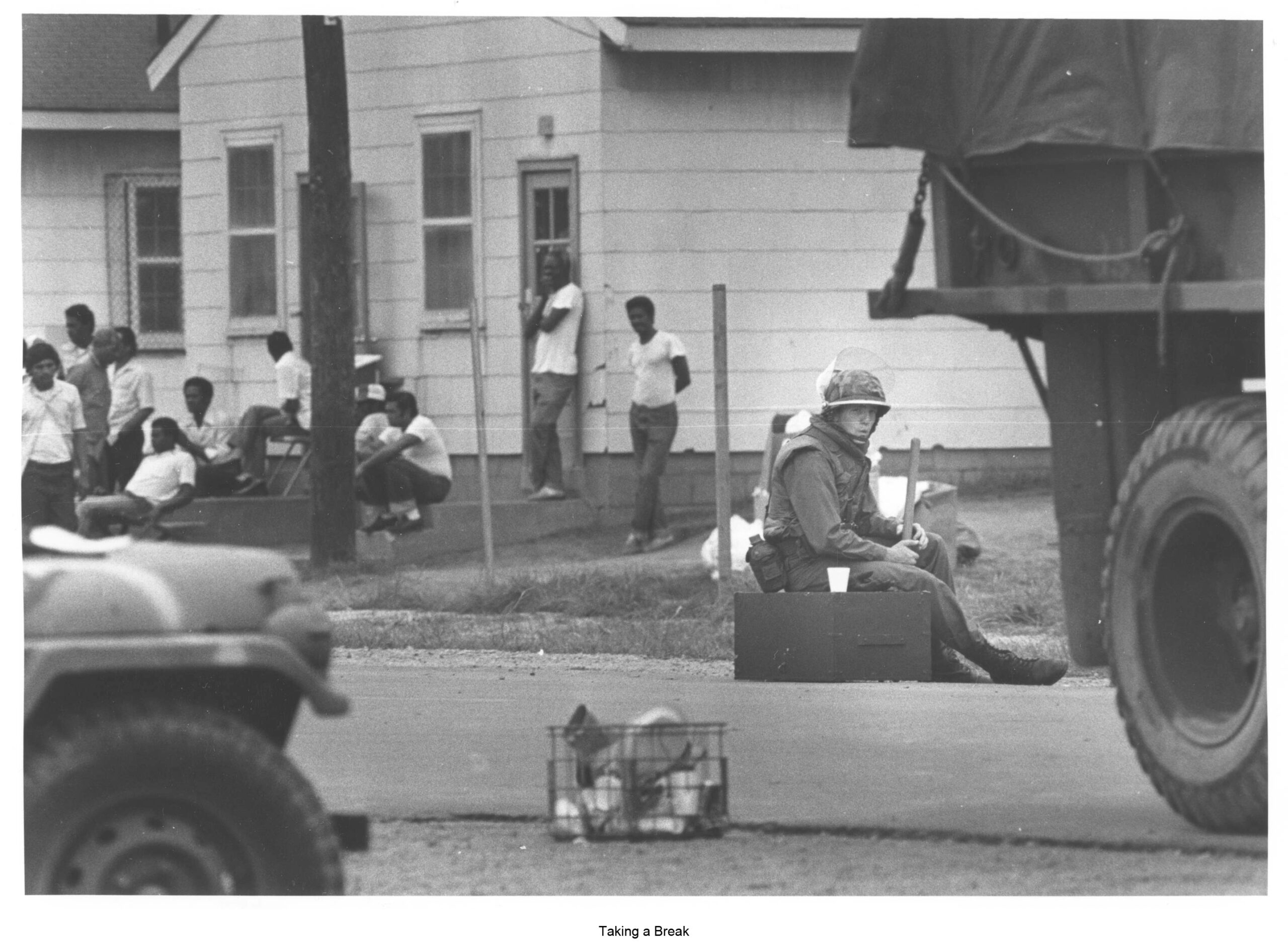 A black and white photograph featuring a soldier with nightstick in hand whisteling as he sits in the streets in front of the single men's compund at Fort McCoy