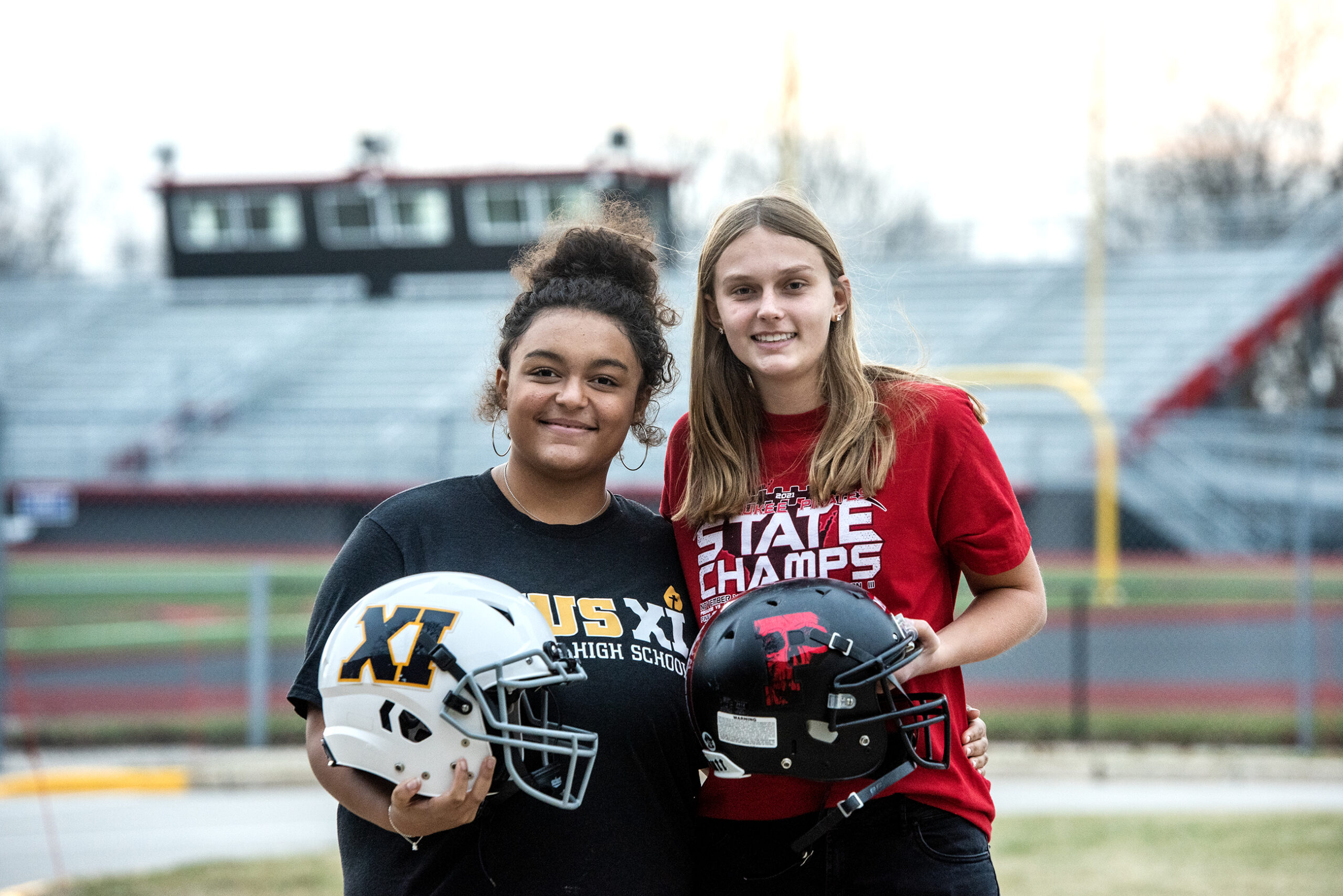 Hannah Peters and Ava Matz stand in front of stadium bleachers and hold their football helmets.