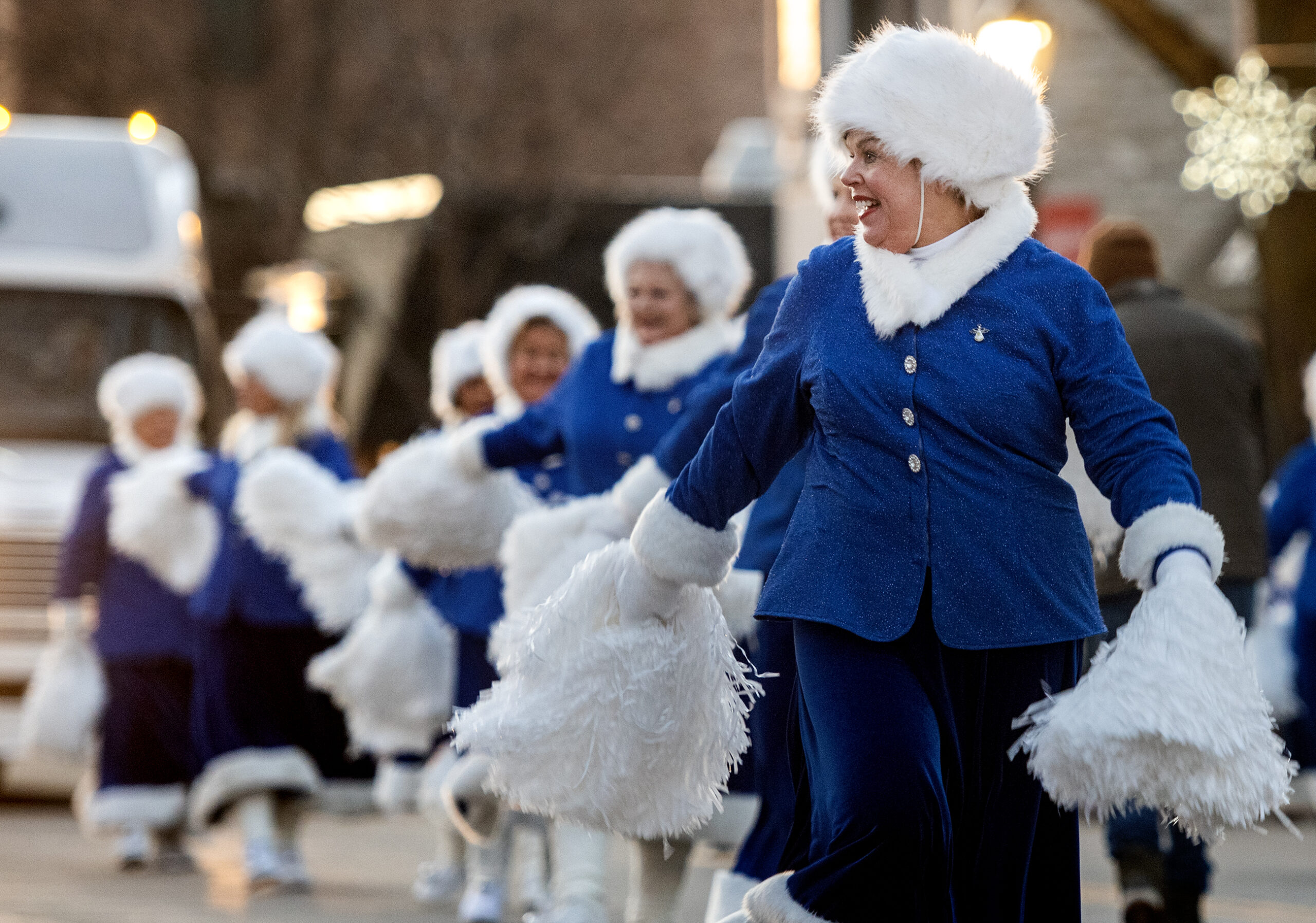 Women in blue coats perform with white poms. They dance together and smile at the crowd.