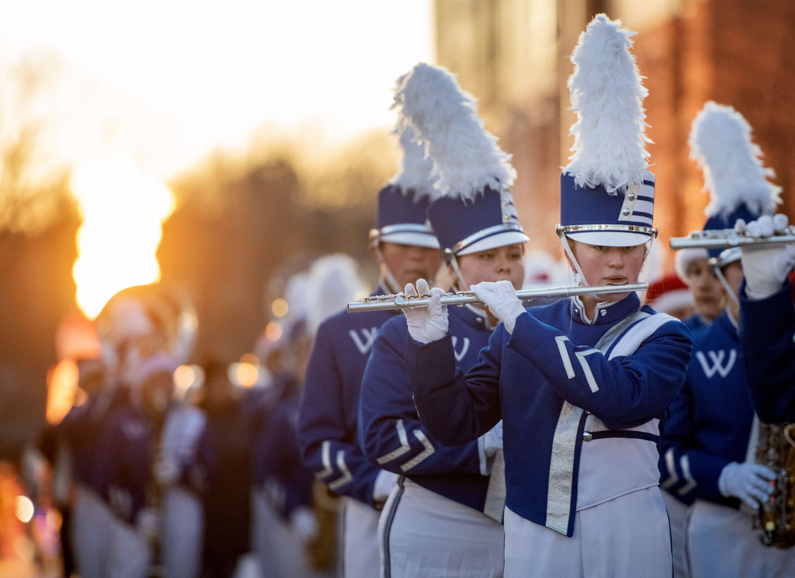 Band members in blue and white uniforms play instruments as the sun sets.