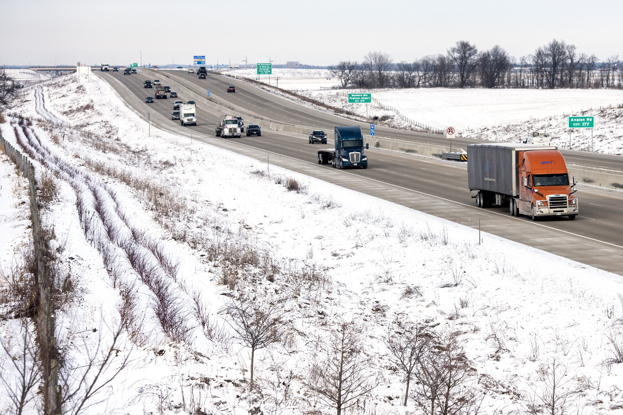Cars and semis drive on an interstate highway past a snowy landscape.