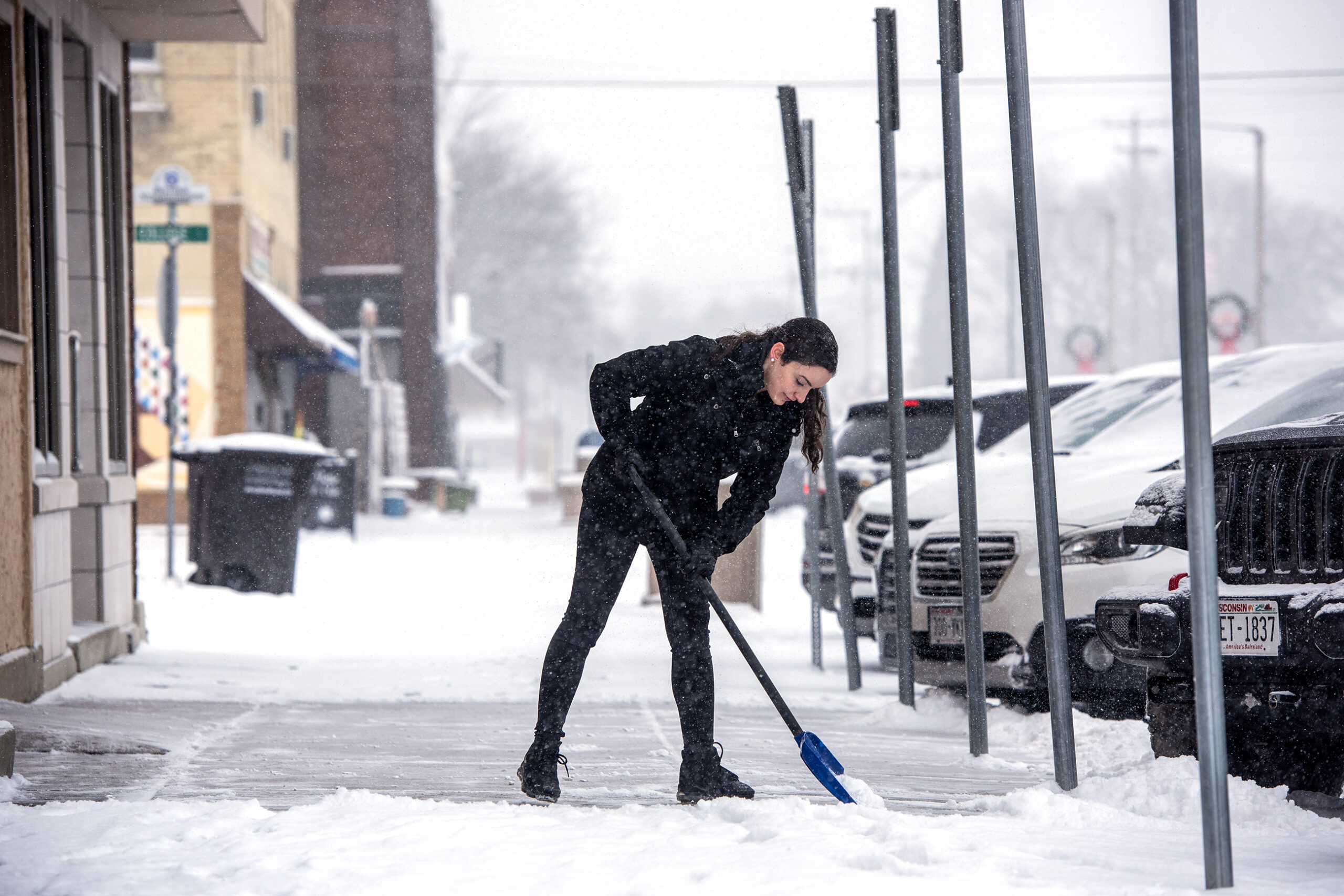 A woman in a coat and boots uses a shovel to clear snow from a downtown sidewalk.