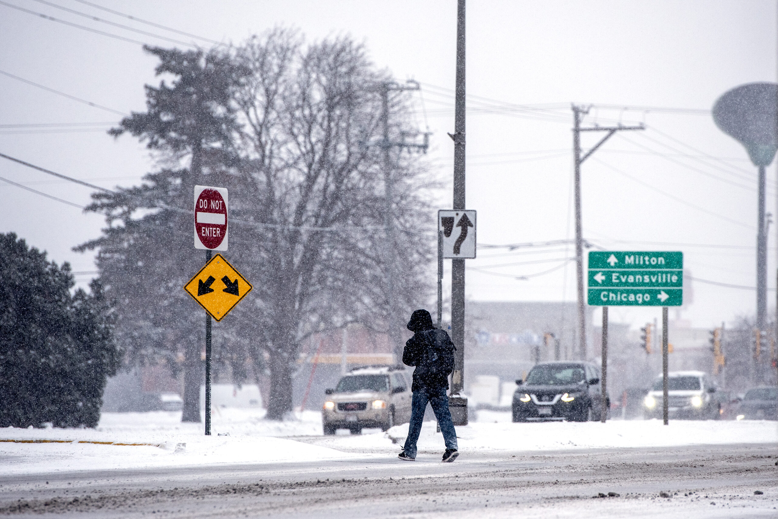 A pedestrian in a black coat and hood approaches the middle of a divided highway in a snow storm.