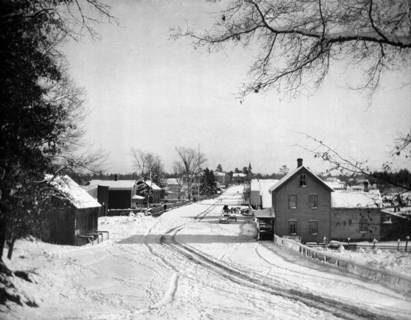 Elevated winter scene of snow-covered road through Keshena, Wisconsin. There are horse-drawn vehicles and pedestrians, and a church is in the background, circa 1902.