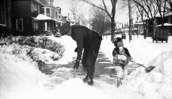 Winter scene with a man and young boy shoveling snow from a sidewalk after a heavy snowfall, circa 1925 in Milwaukee.