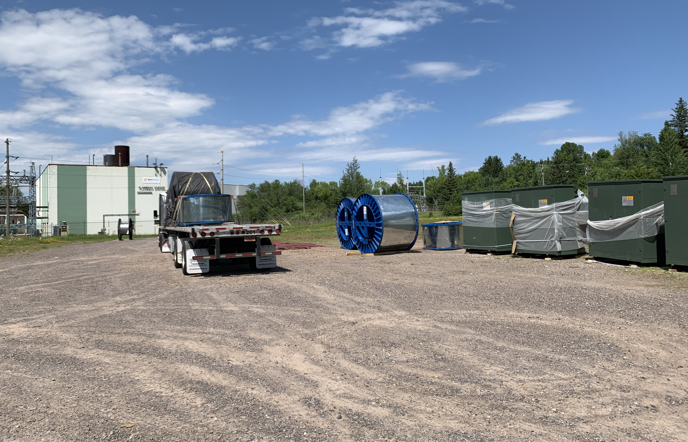 An empty semi truck is parked near some large equipment next to a shed on gravel