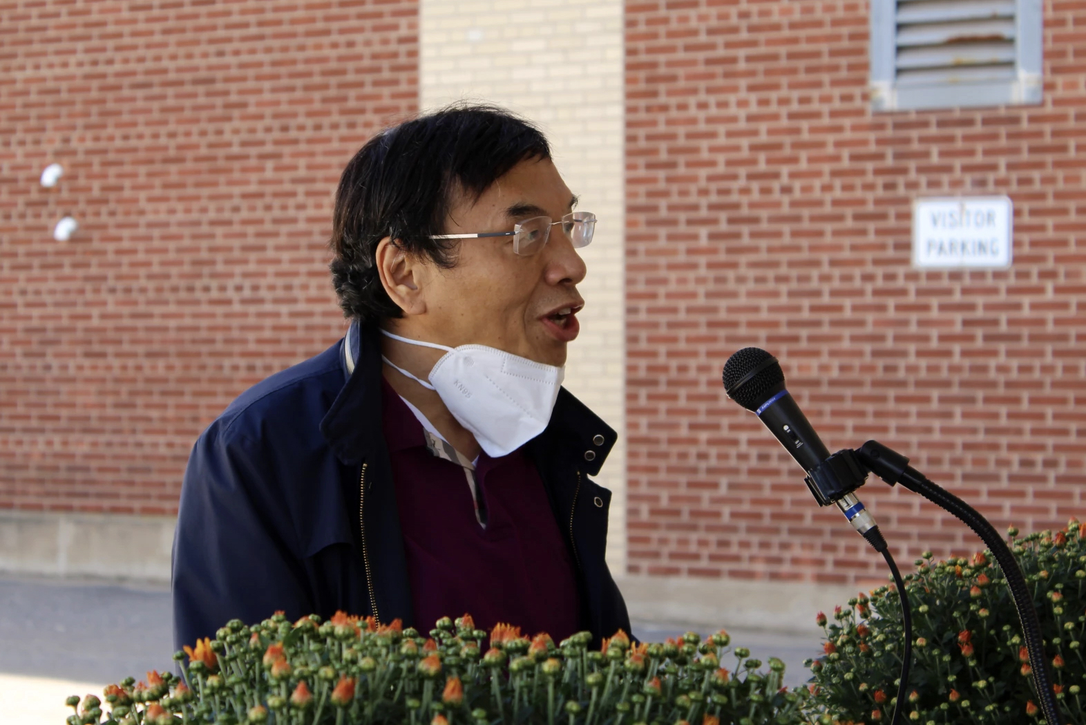 Yong Liu speaks at a podium into a microphone, with his face mask on his chin