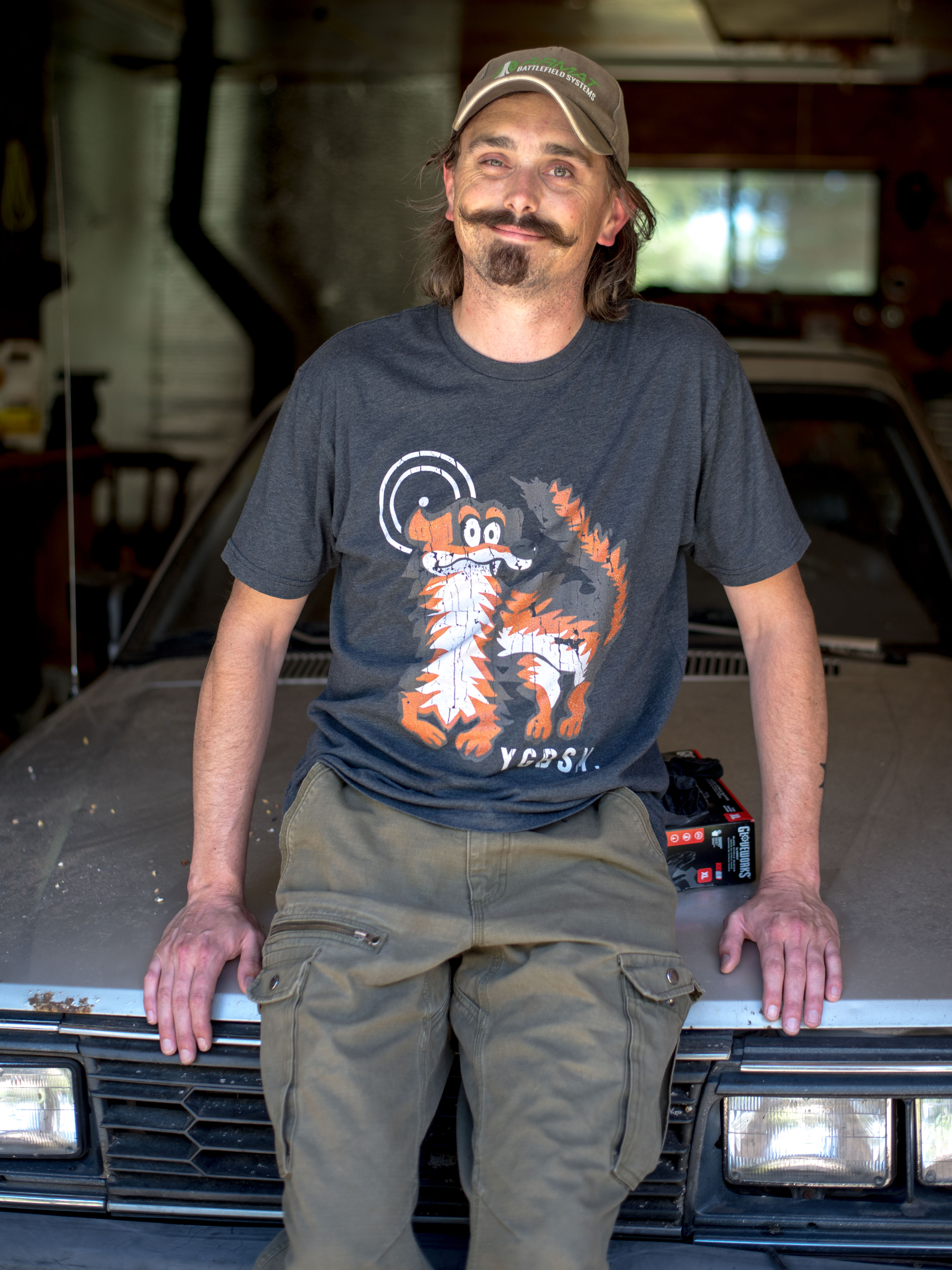 Zach Skrede poses for a portrait. He is leaning on a hood of a car and smiling