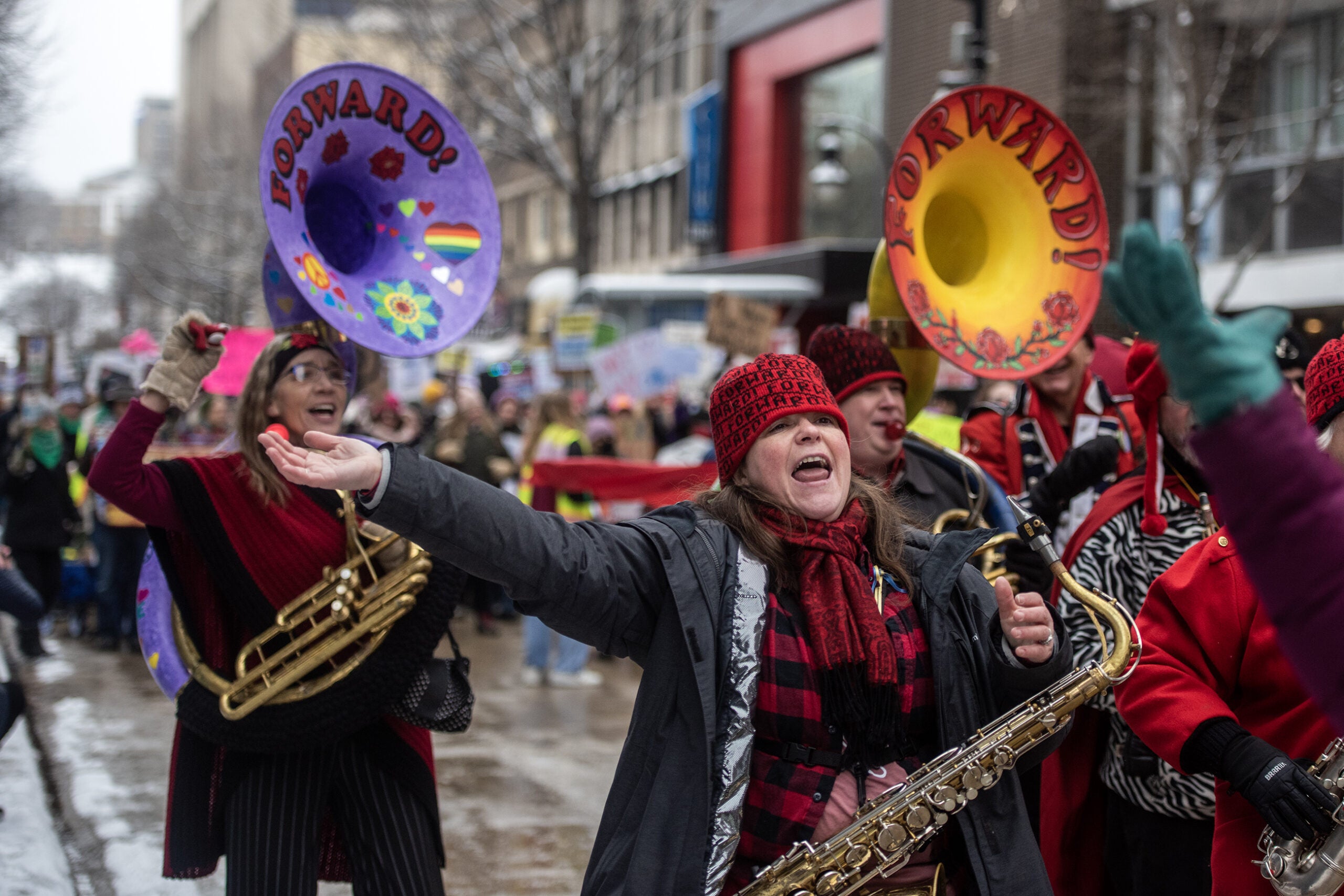 A saxophone player holds out her arm and sings with other band members while marching outside.