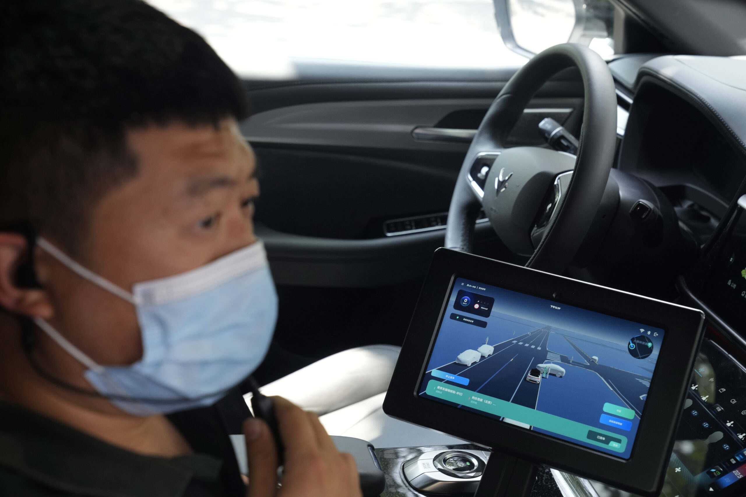 A man with a covid mask on sits and works on a screen in a car. The screen has a map on it.