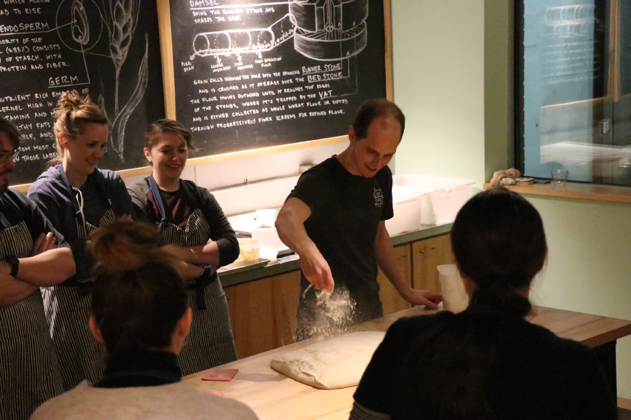 People in Madison Sourdough watch as a baker throws flour on some dough