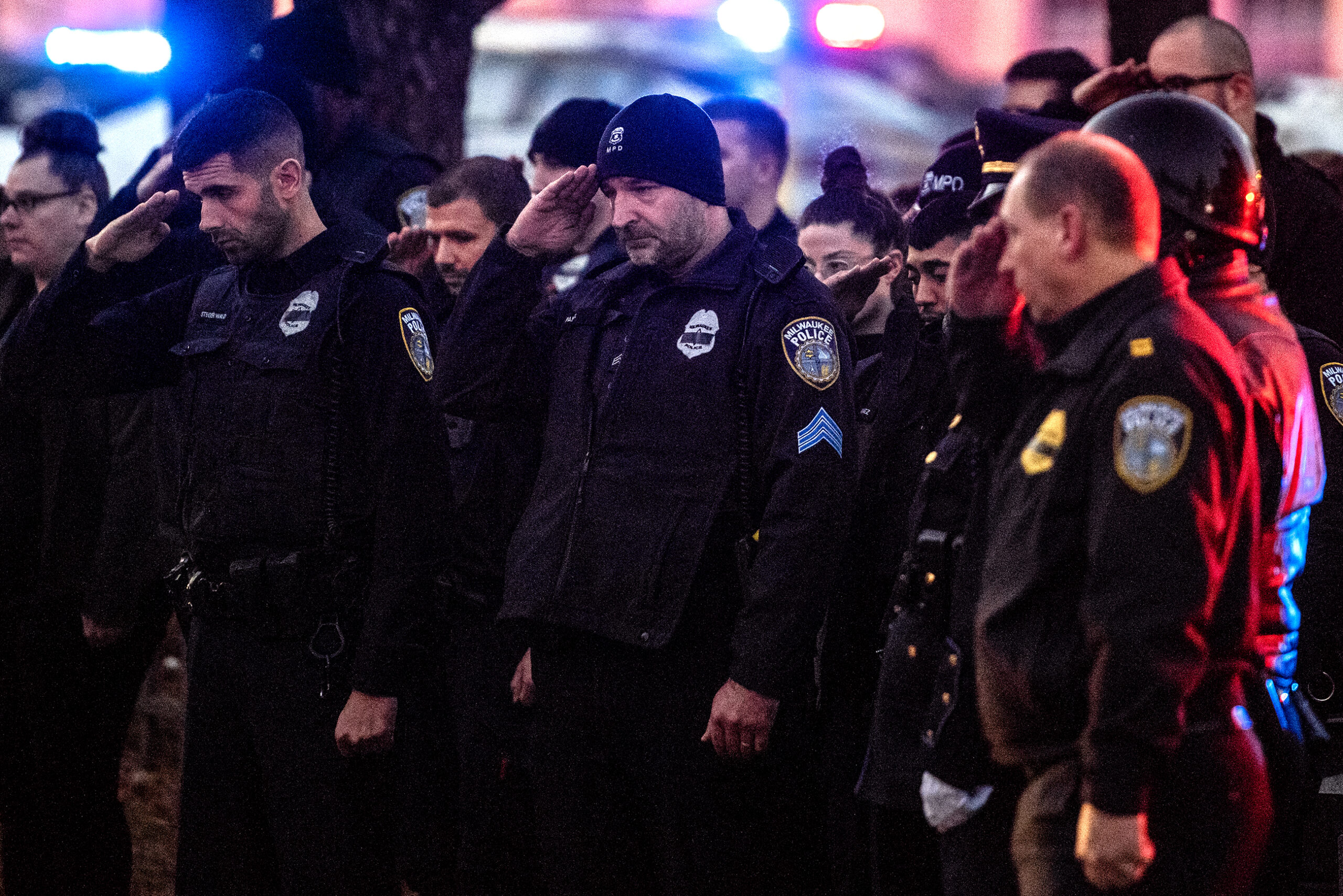 A line of police officers in uniform hold their hands up to salute. Police lights can be seen in the background.