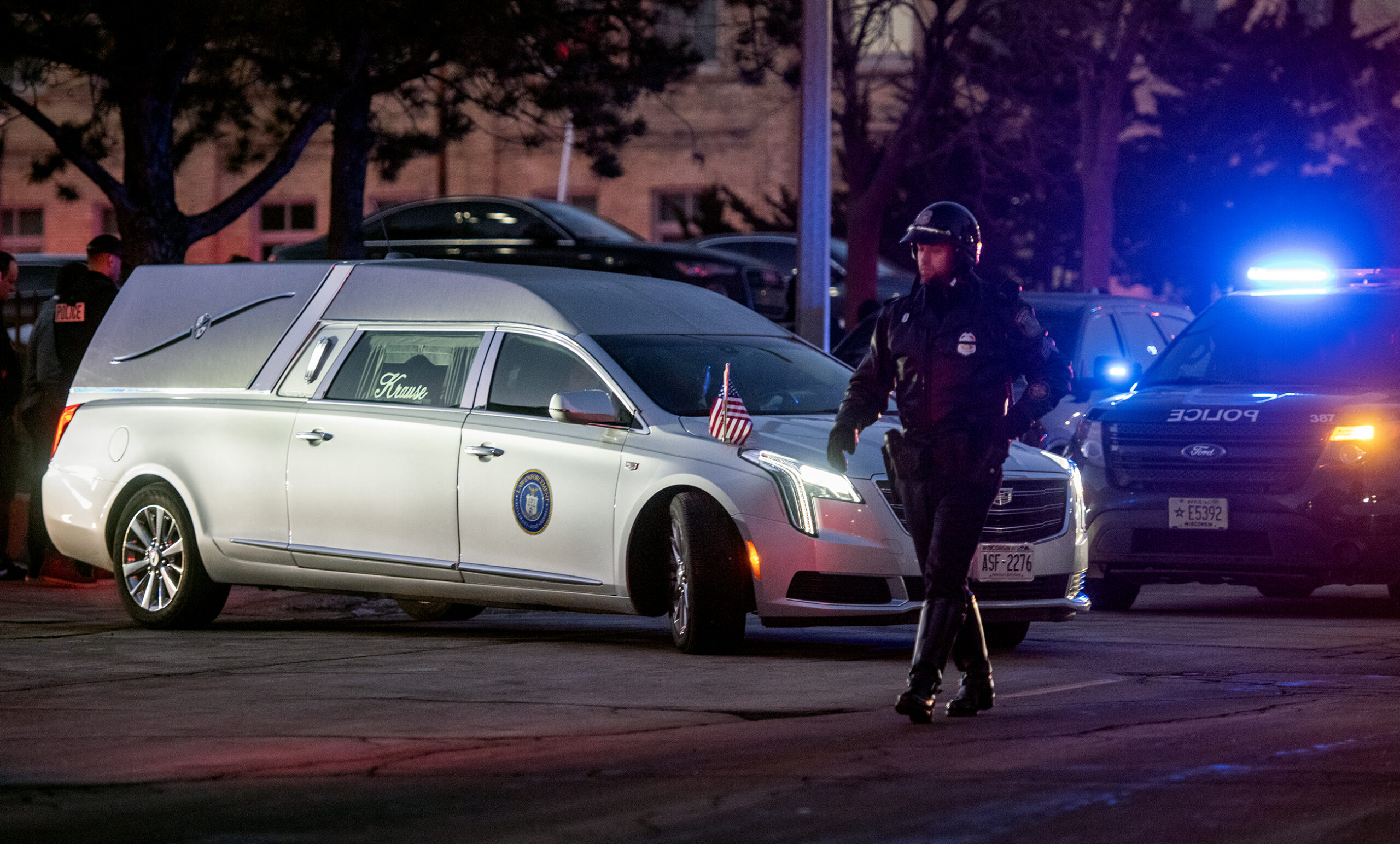 A silver hearse pulls out in front of a police vehicle with lights on. A police officer in uniform walks nearby.