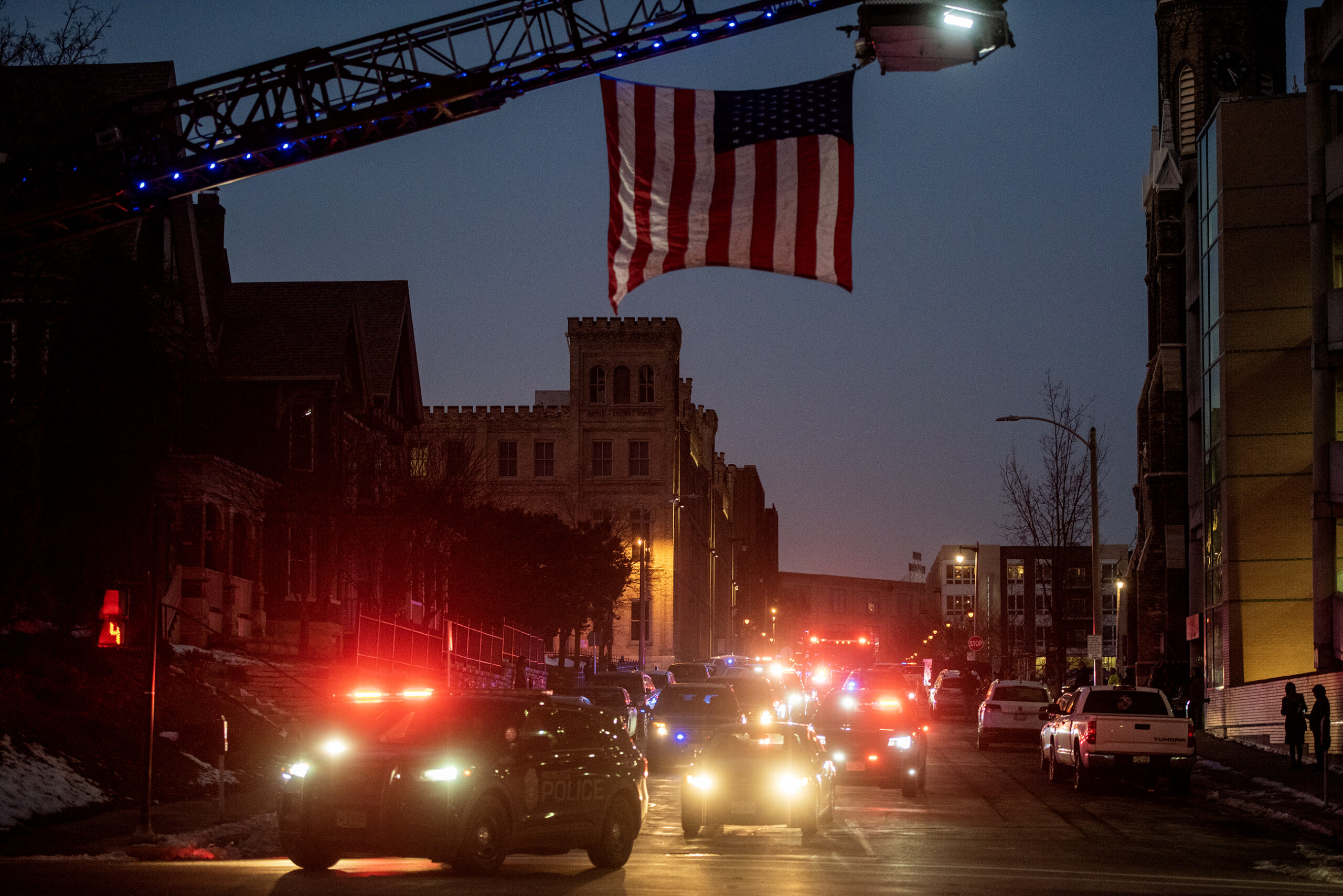 A U.S. flag hangs from a fire truck above the procession of police cars with lights on.
