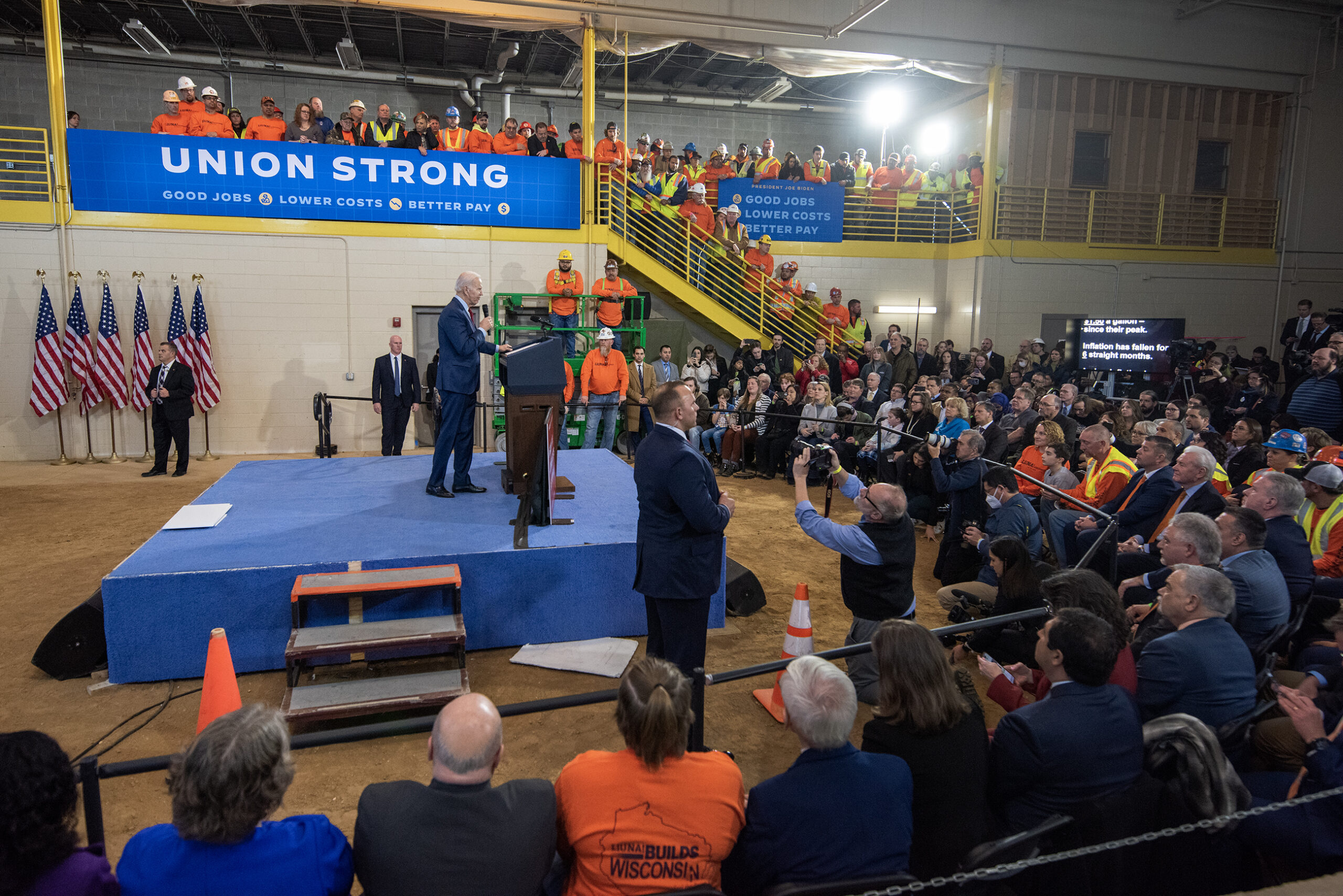 President Joe Biden is seen in the middle of a crowded room. A sign in the background says 