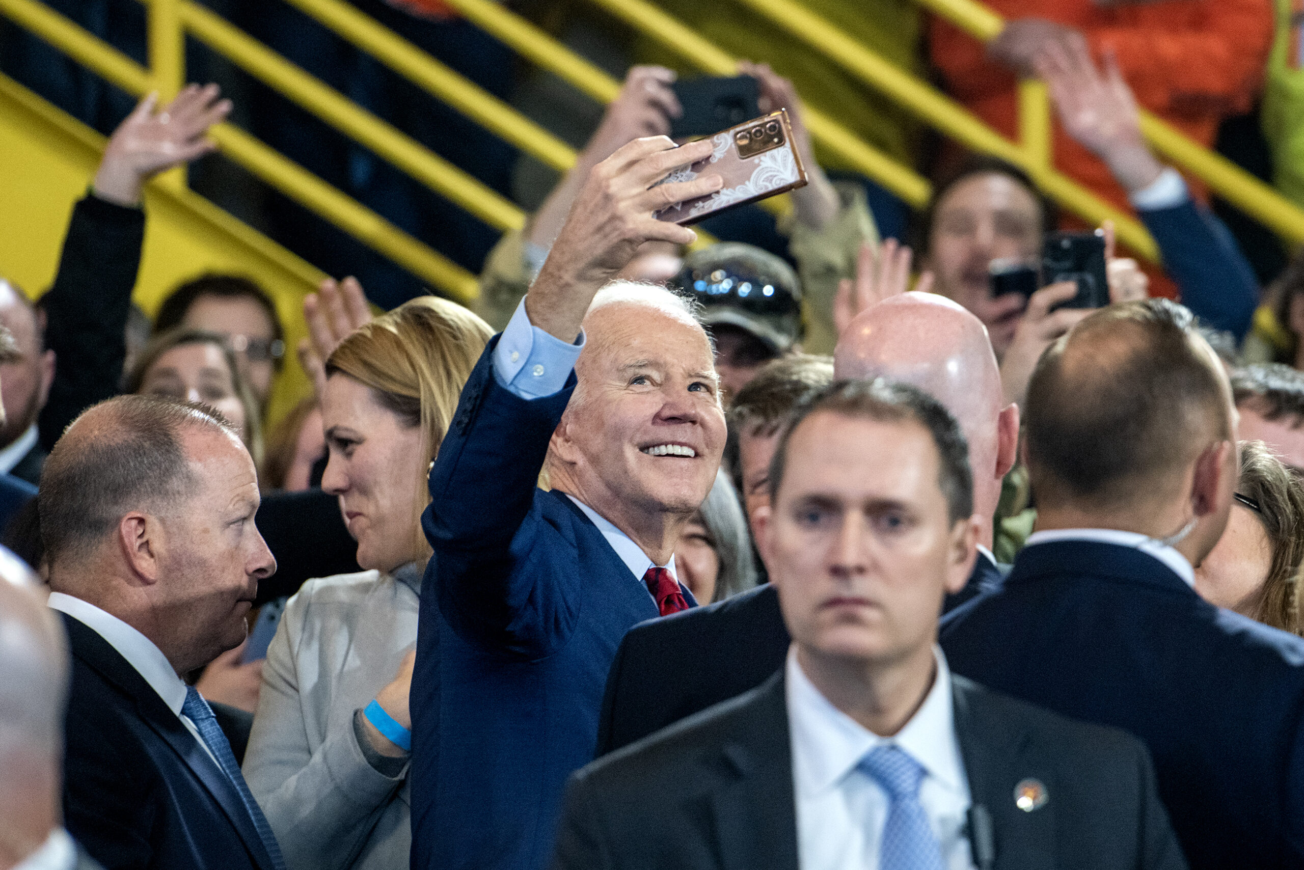 President Joe Biden holds a phone in the air while smiling and taking a selfie.