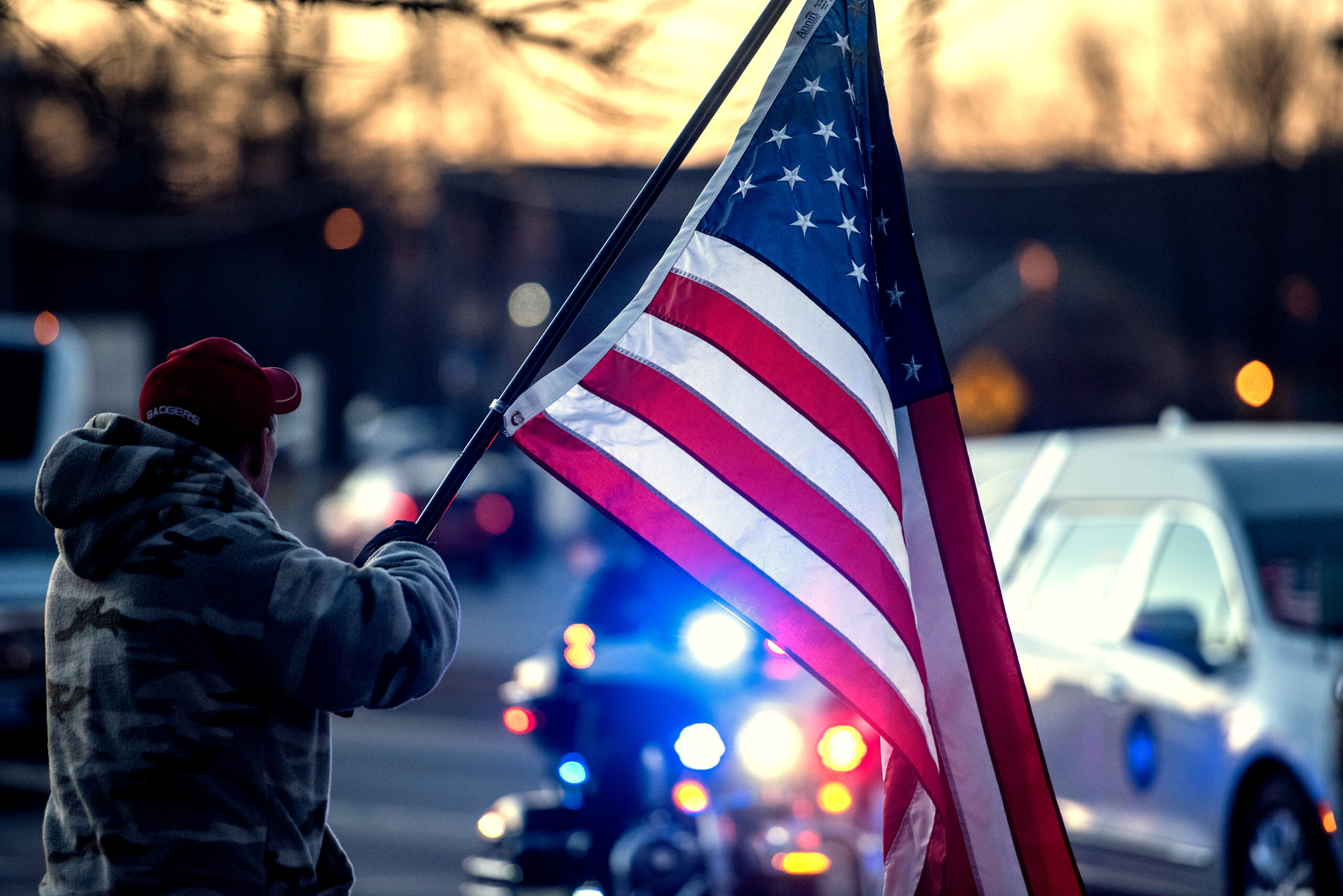 The stripes of a U.S. flag are illuminated by blue and red police lights. A white hearse passes by in the background.