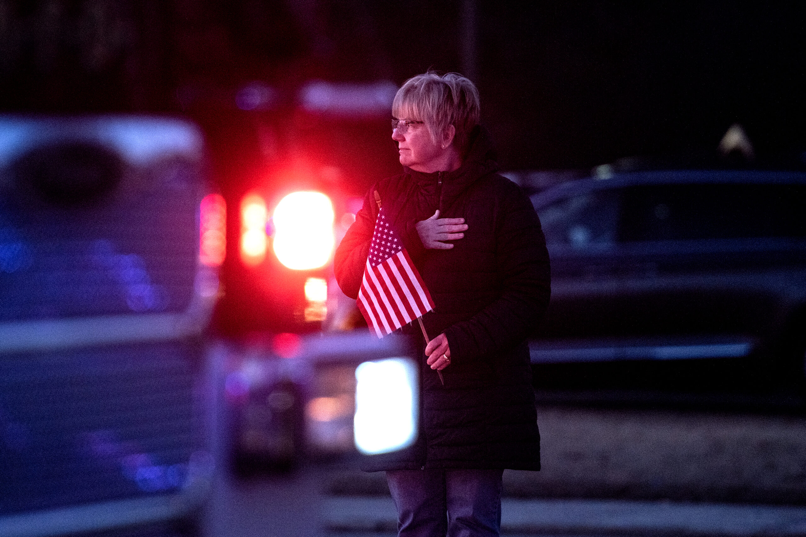 Red light from a fire truck shines into the dark as a woman holding a small U.S. flag watches with her hand on her heart.
