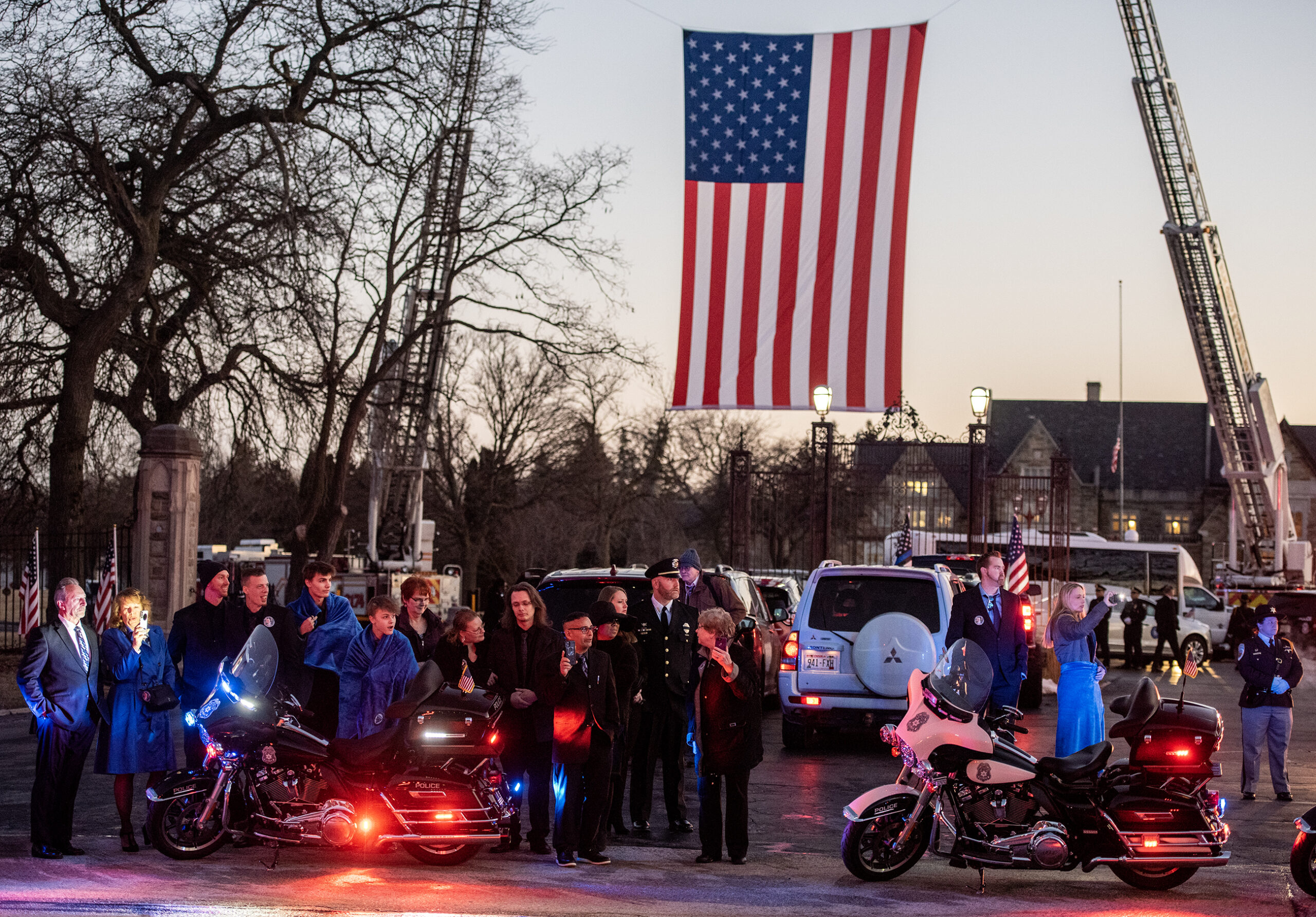 A large U.S. flag is displayed at a cemetery. People gather to watch the procession.