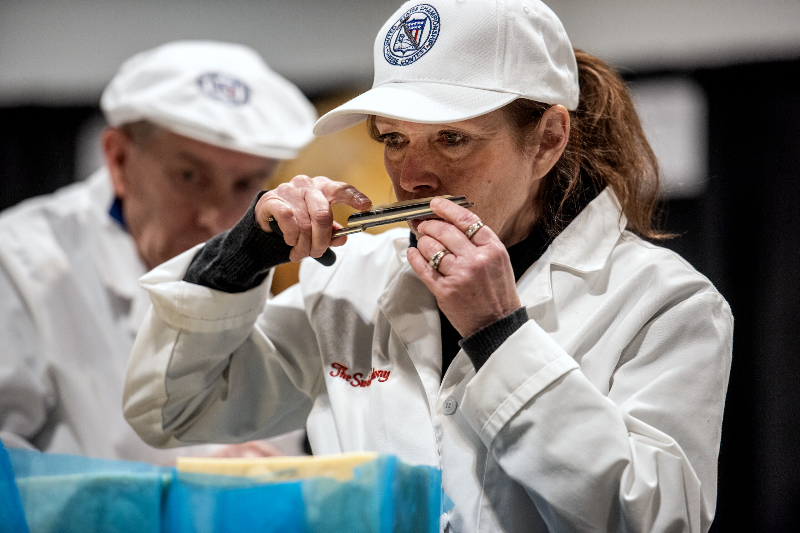 A woman in a white coat and hat sniffs a piece of cheese.