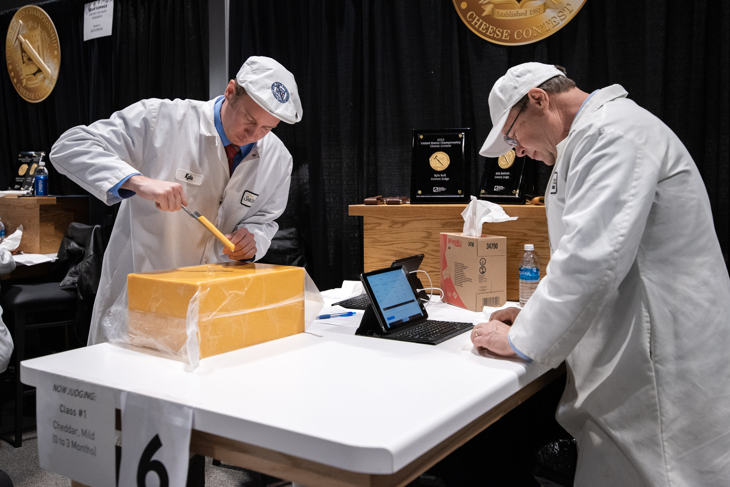 Two judges stand at a table around a large block of yellow cheese.