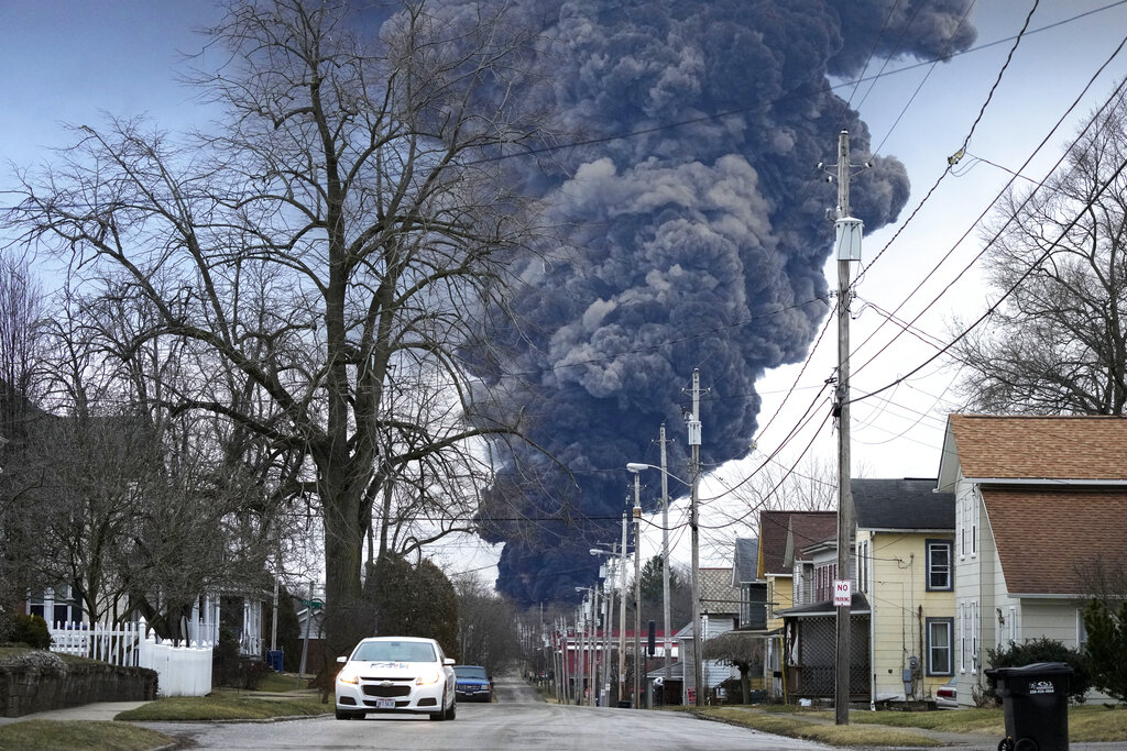 A black plume of smoke rises in the sky above a residental neighborhood.