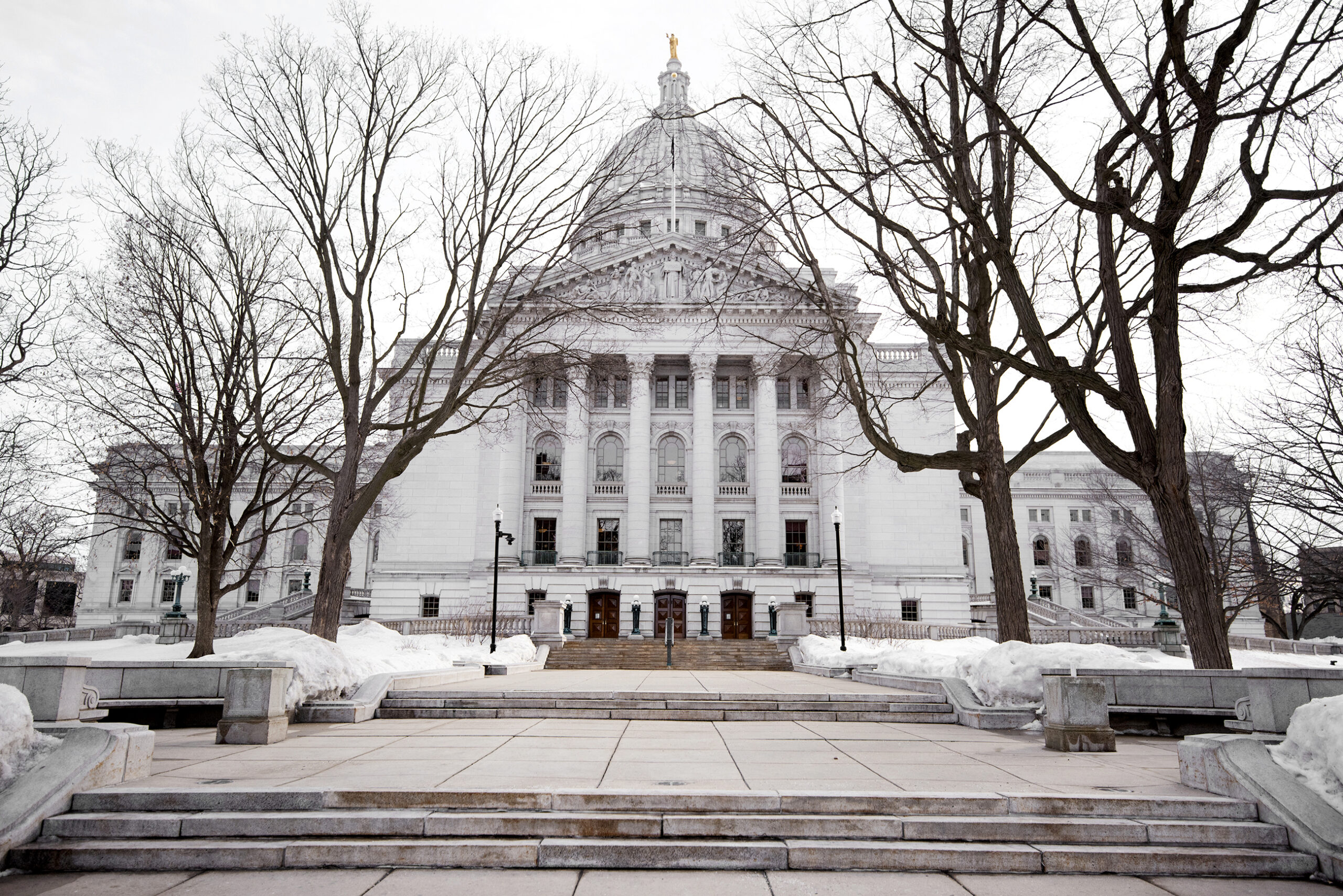 A gray sky backdrops the Wisconsin State Capitol. Snow is on the ground and there are no leaves on the trees.