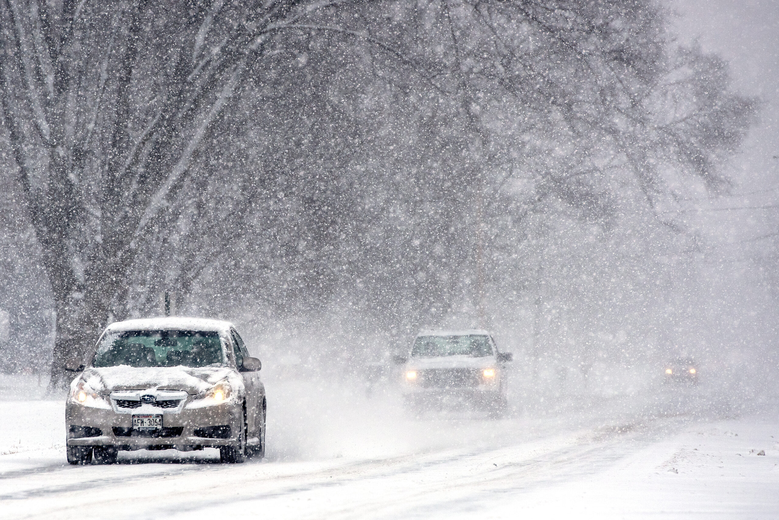 Three vehicles drive on a snow covered road during snowy conditions. The two cars in the distance can hardly be seen.