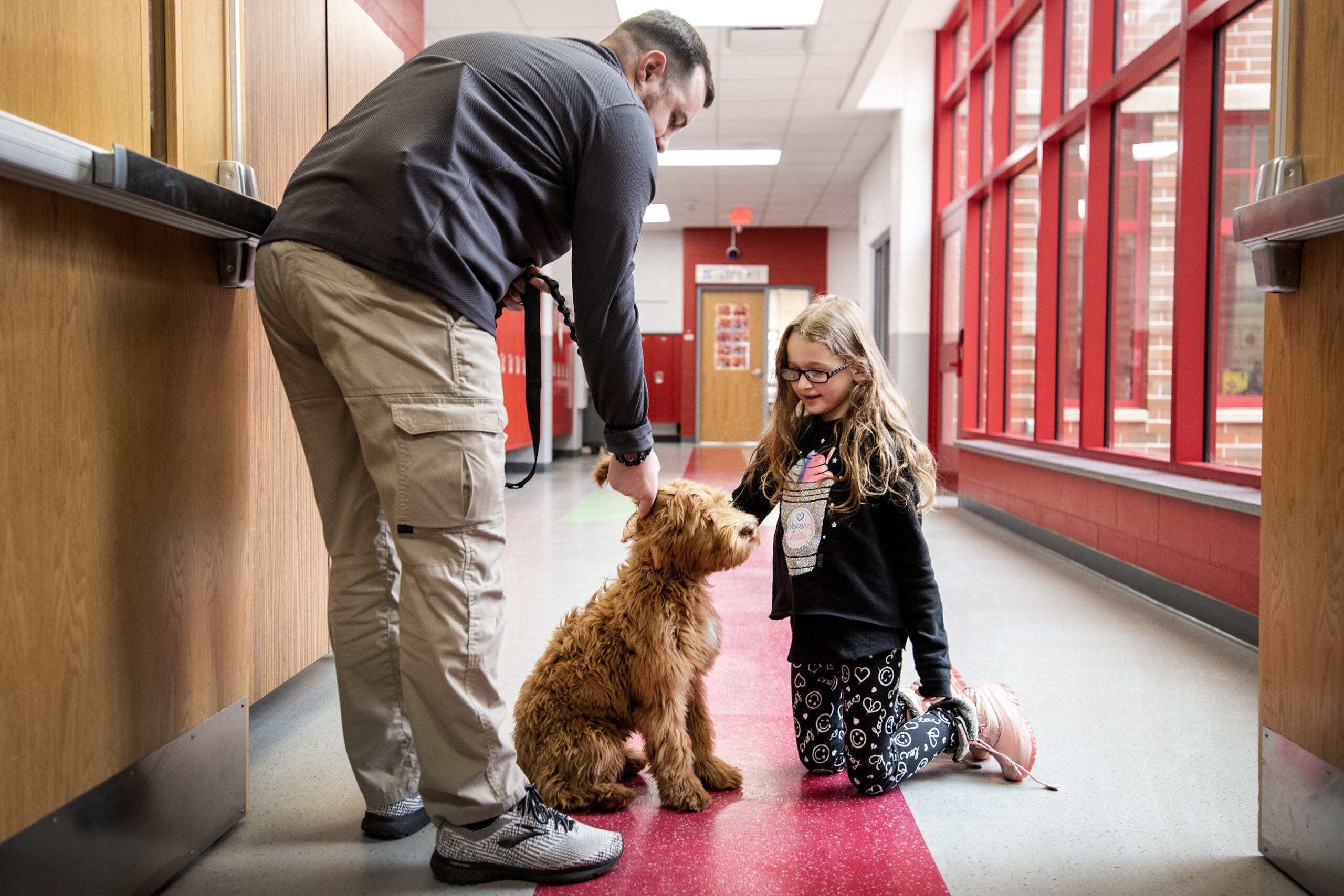 A girl kneels down to pet a brown dog in a school hallway.