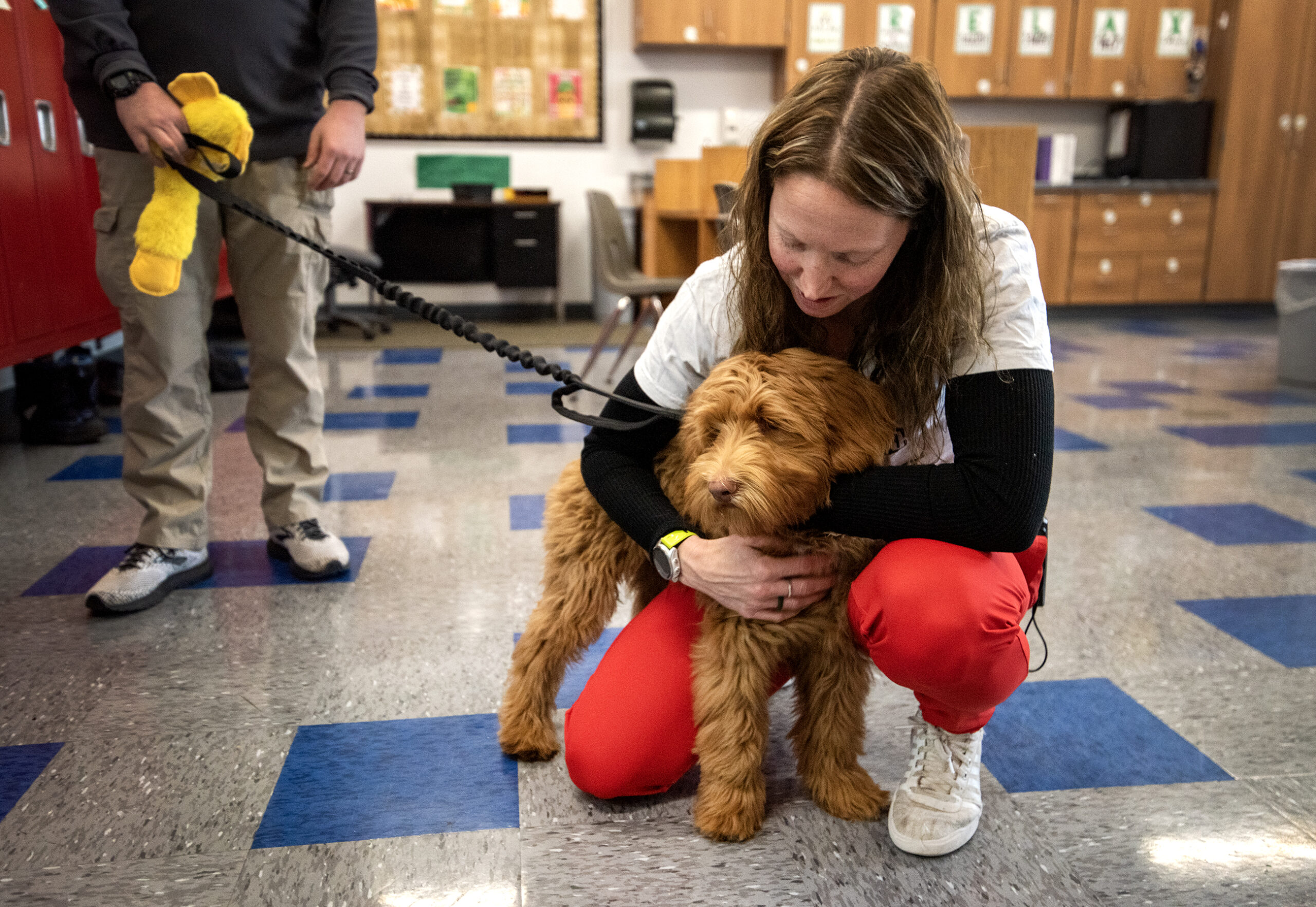 A teacher kneels to hug a fuzzy brown dog in a classroom.