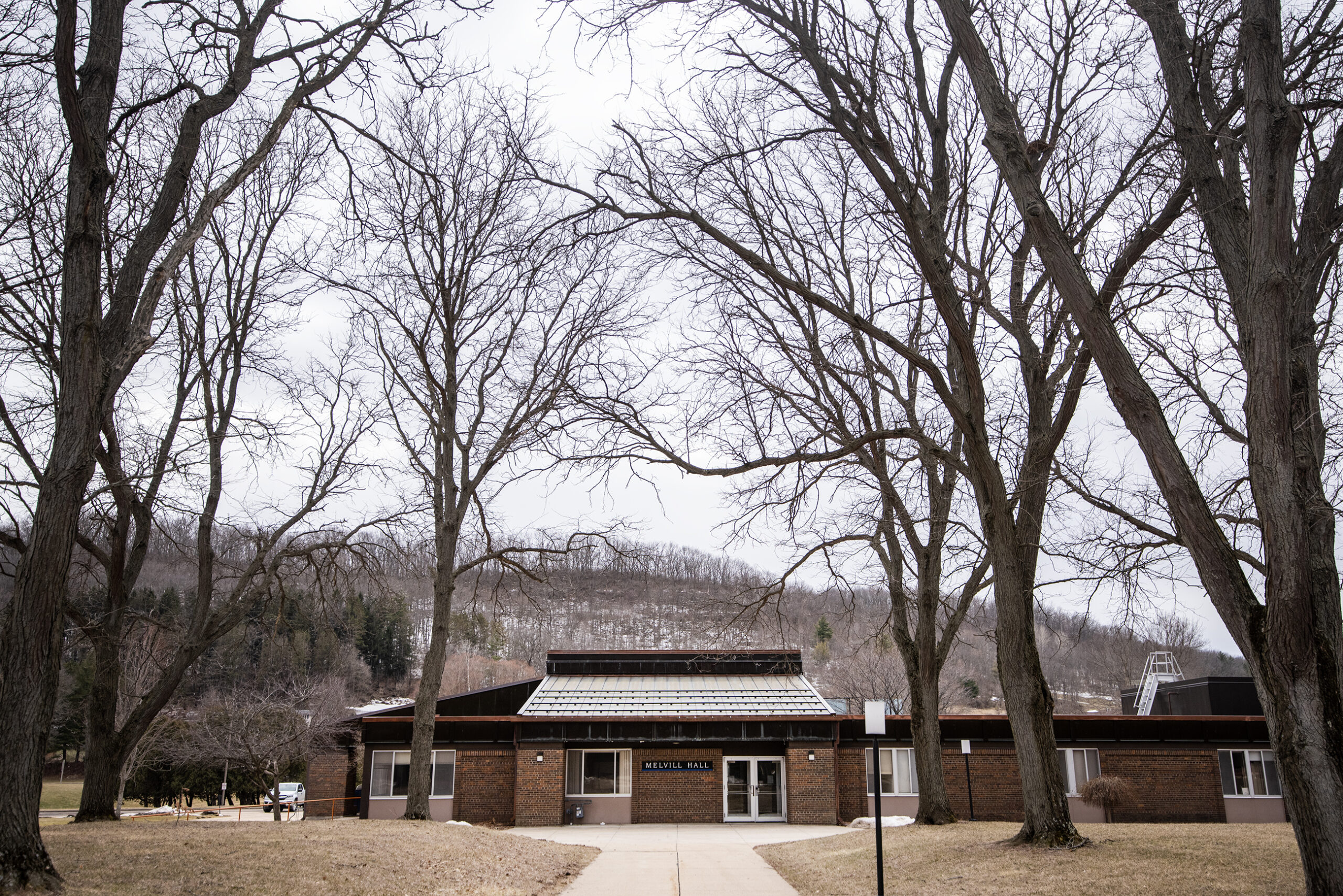 Bare trees stand tall above a small brown brick building. Hills can be seen in the background.