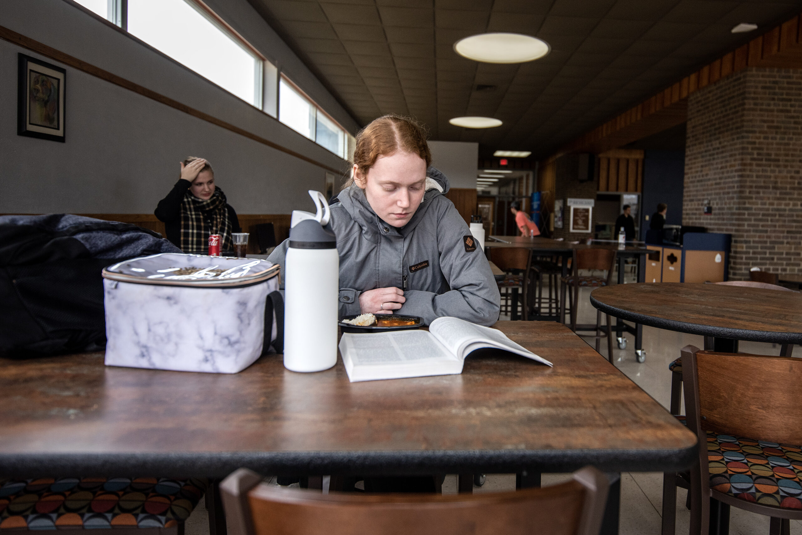 A student sits at a table and reads a book. A lunchbox and water bottle are next to her.
