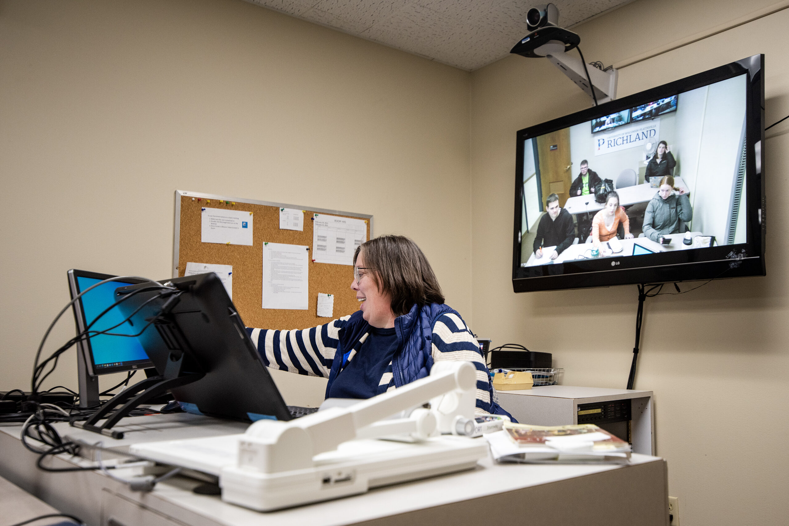 A teacher sits in front of computer screens as classrooms are streamed behind her on tv screens.