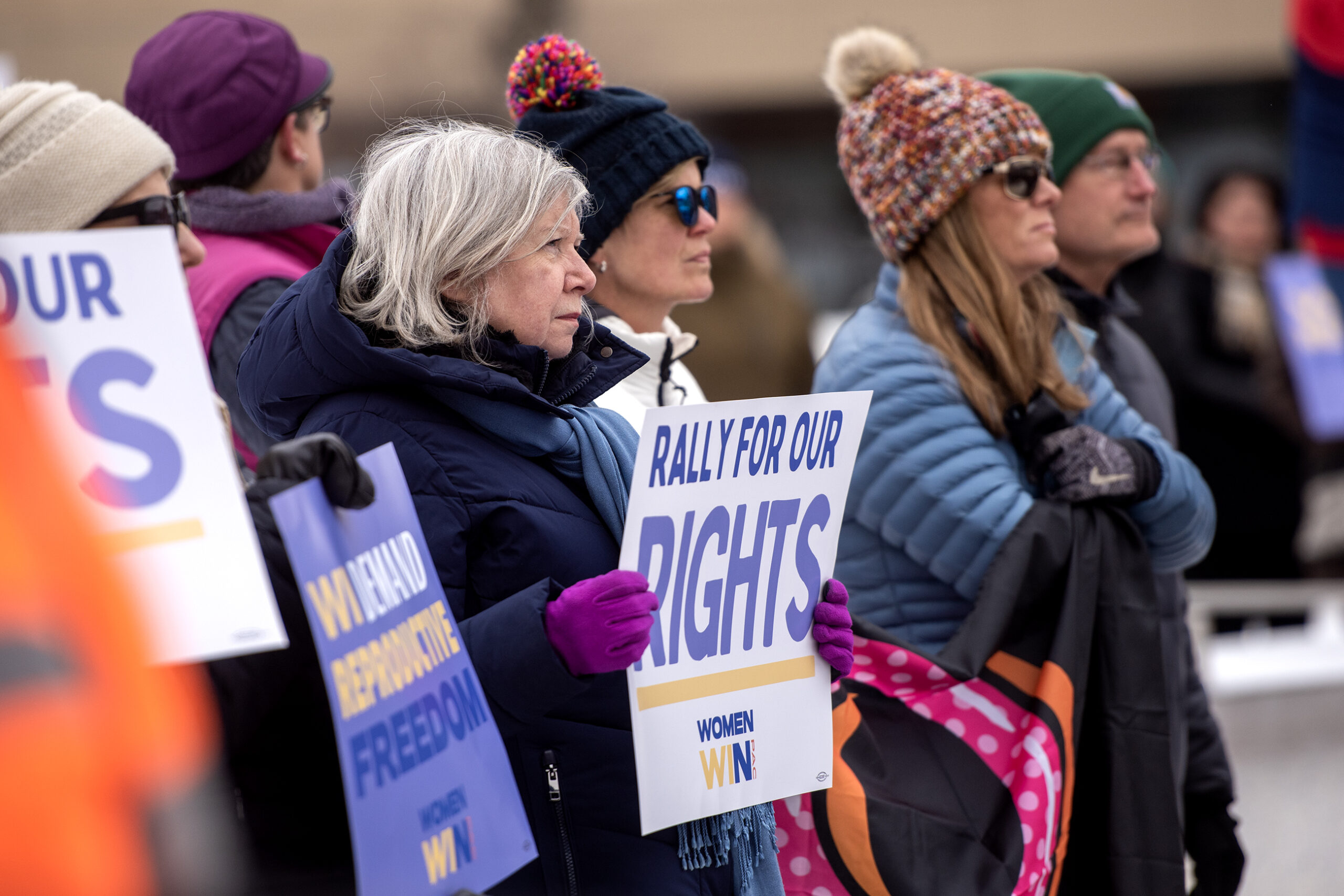 People stand together in winter clothing as they hold signs at a rally.