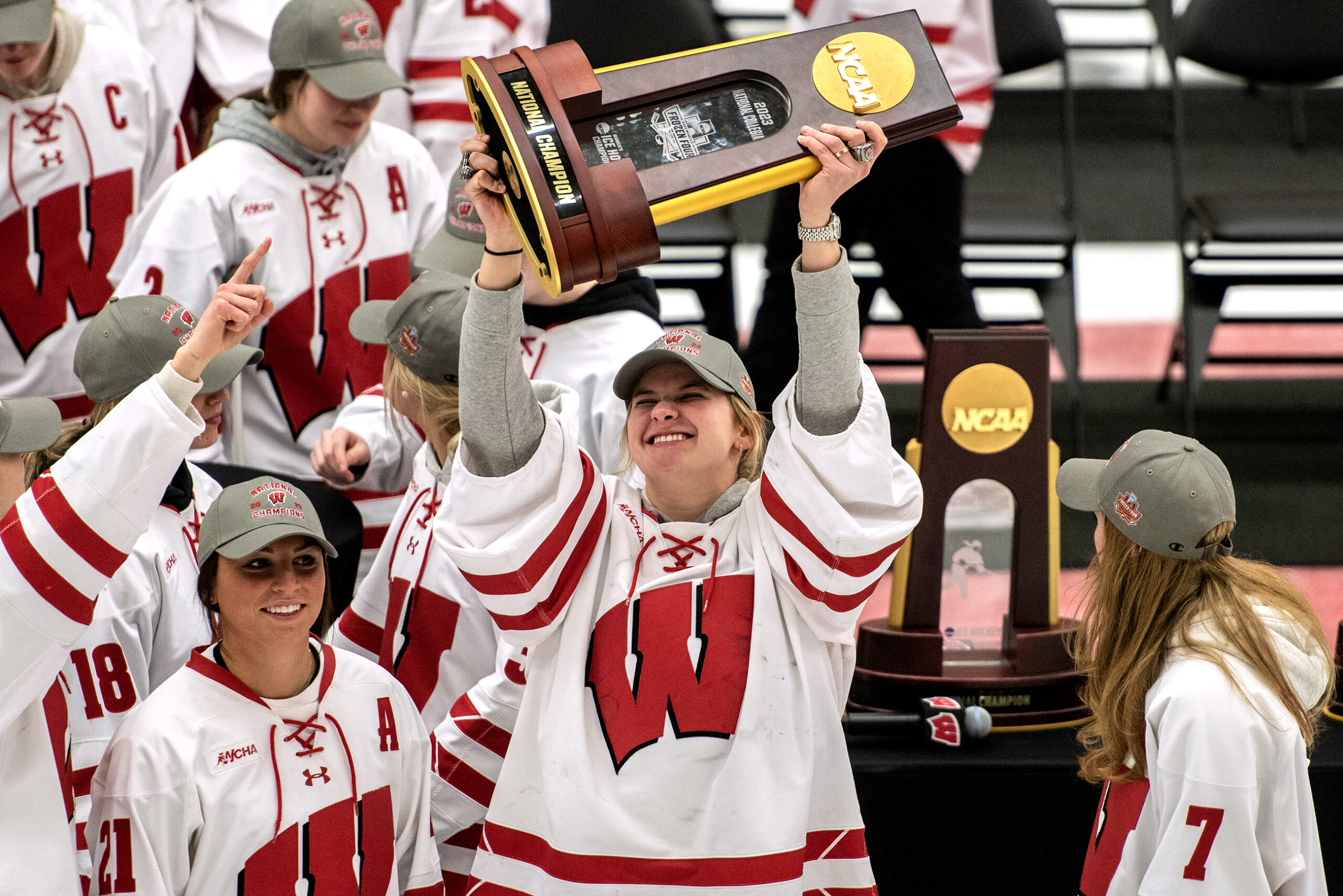 A hockey player in a Wisconsin jersey lifts a wooden trophy with gold details above her head as she smiles.