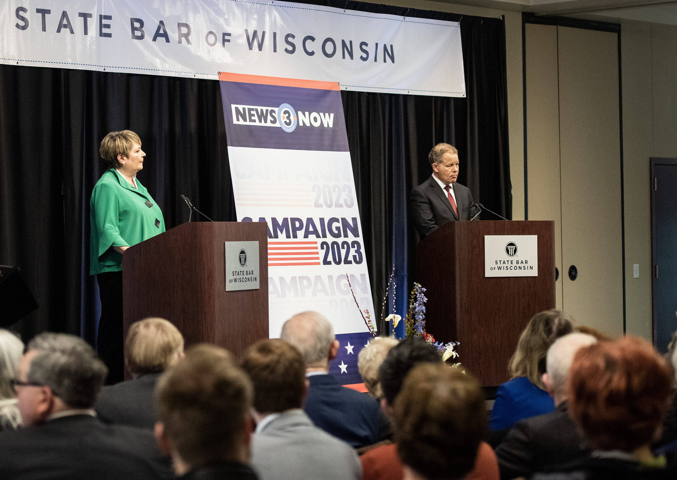 The two candidates stand at podiums on stage. A sign for the State Bar of Wisconsin hangs behind them.