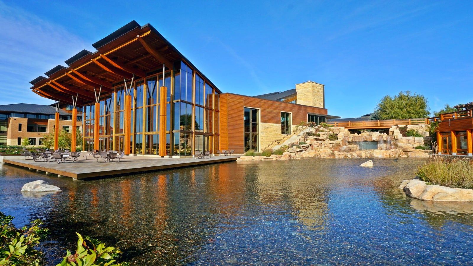 Modern building with large glass windows and wooden beams sits beside a pond with rocks and a waterfall, under a clear blue sky.