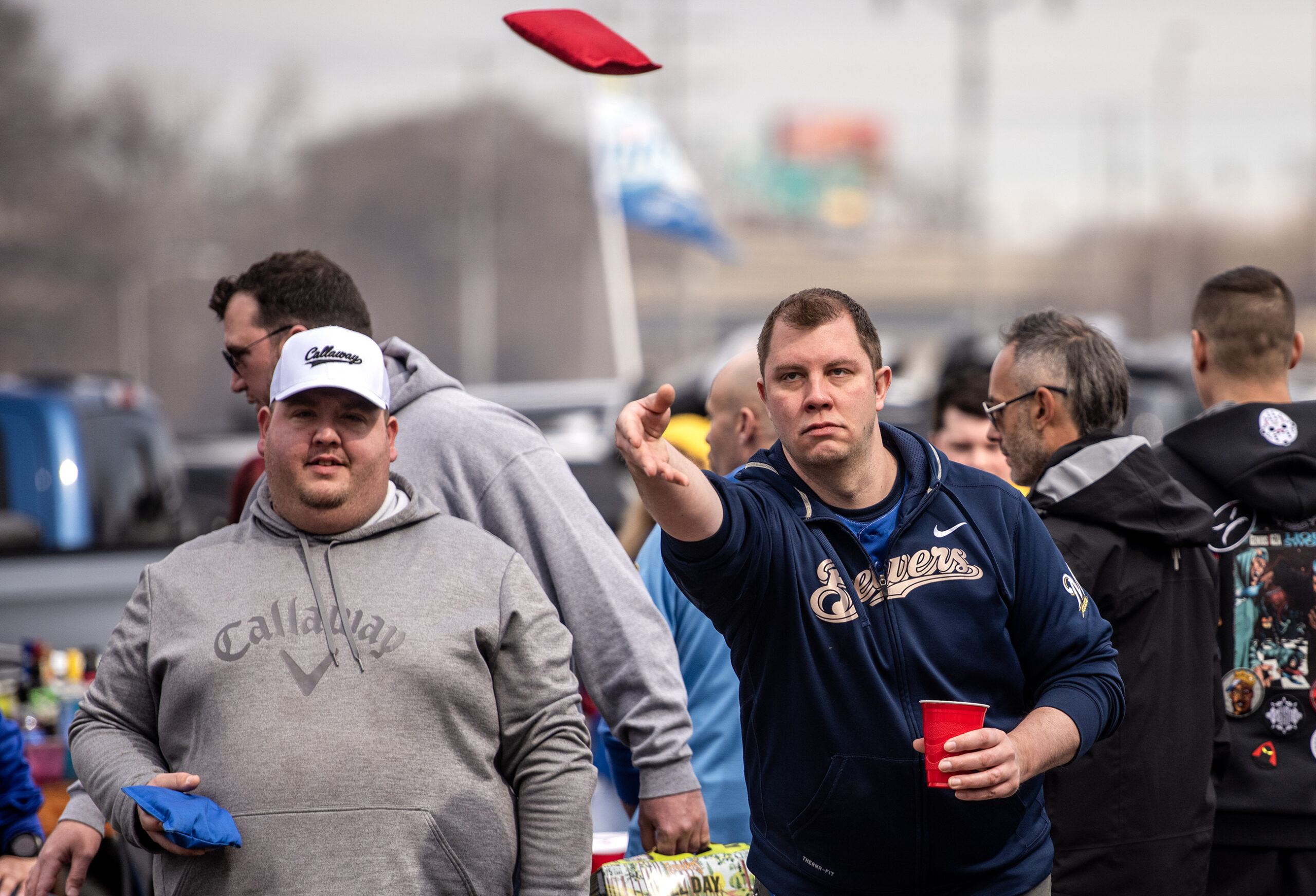 A man throws a red beanbag in the air while playing a game of bags outside as crowds of tailgaters stand around him.