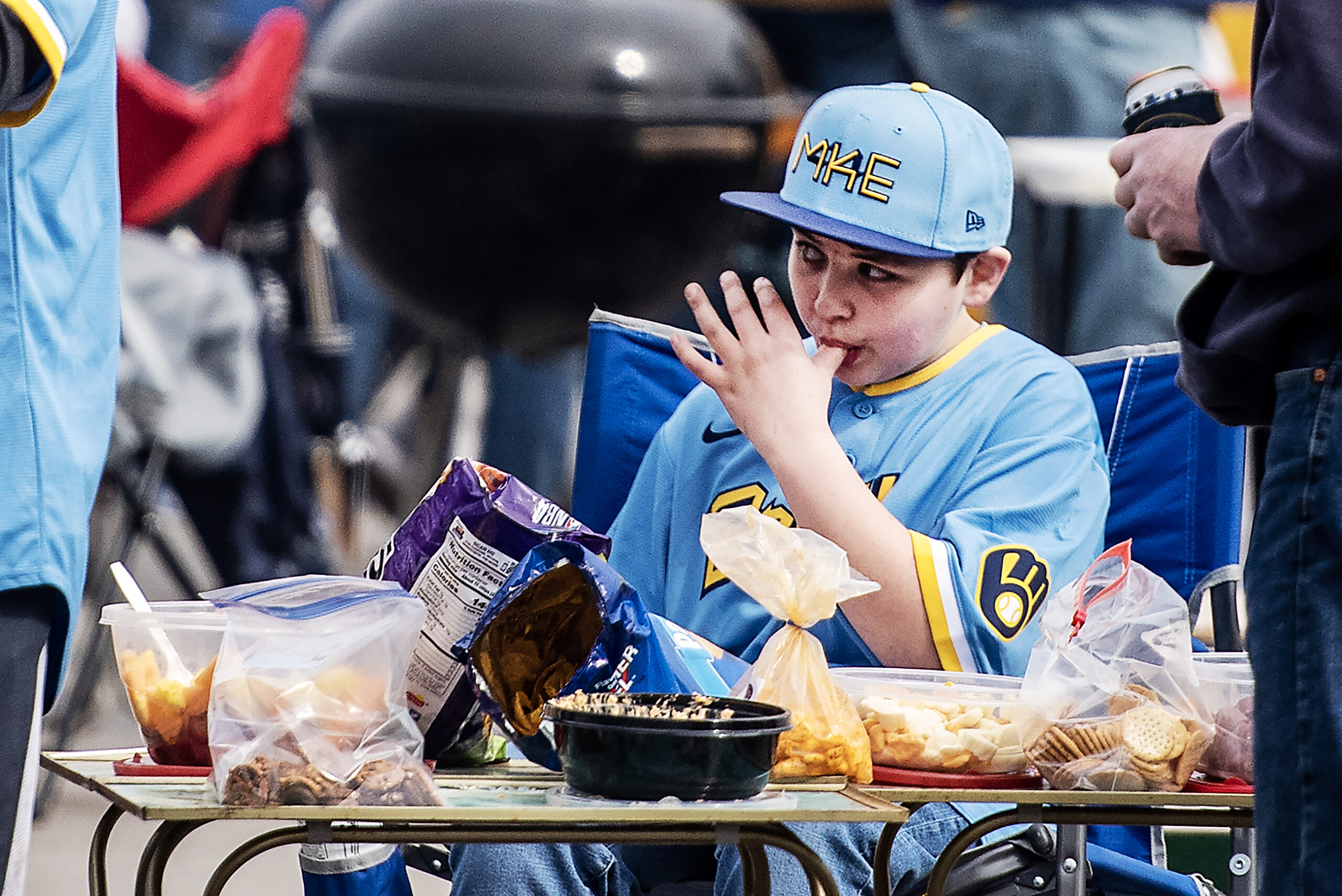 A boy in a blue Brewers jersey and baseball cap licks his fingers while eating tailgate snacks.