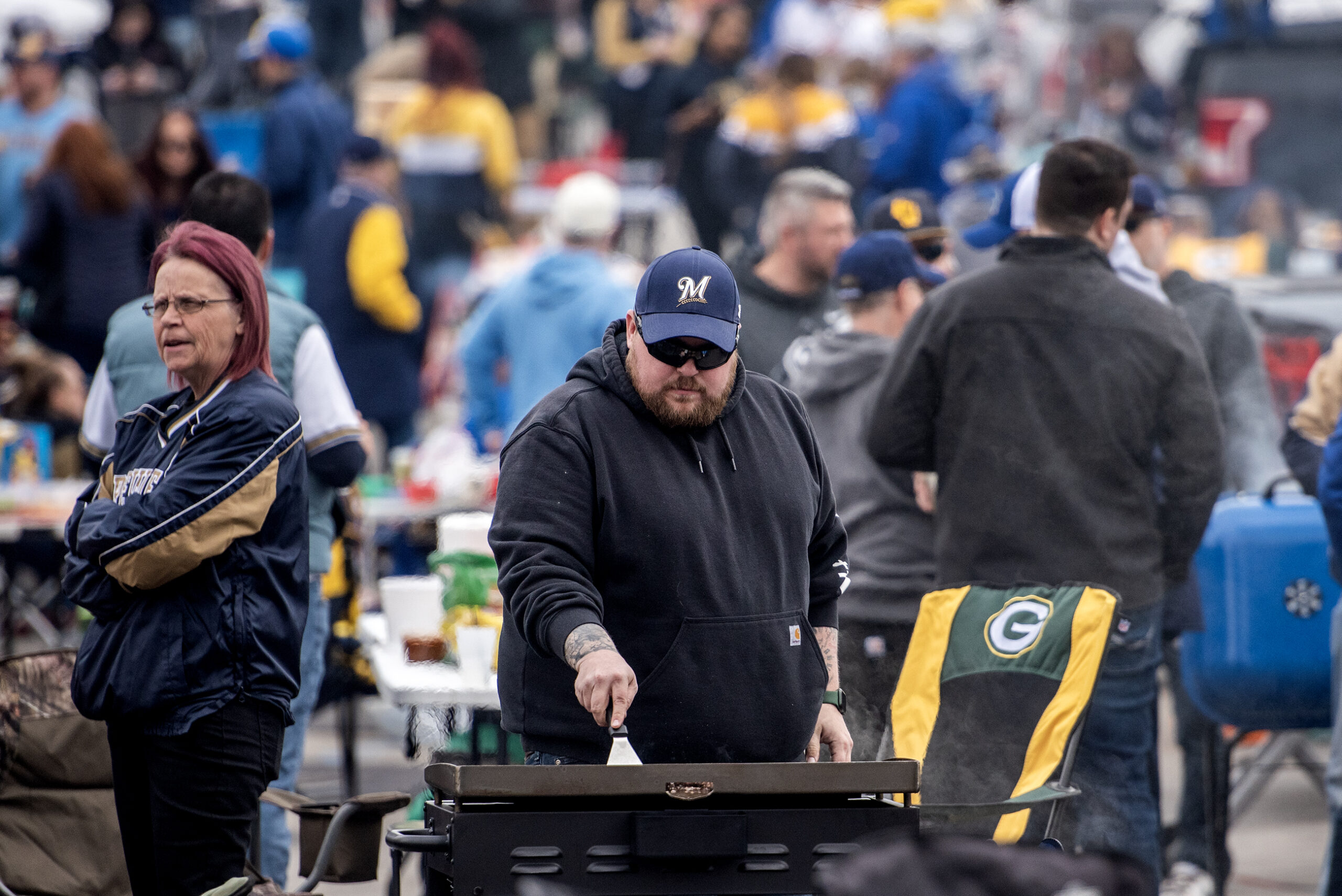 A man uses a spatula to flip burgers on a grill at a tailgate.