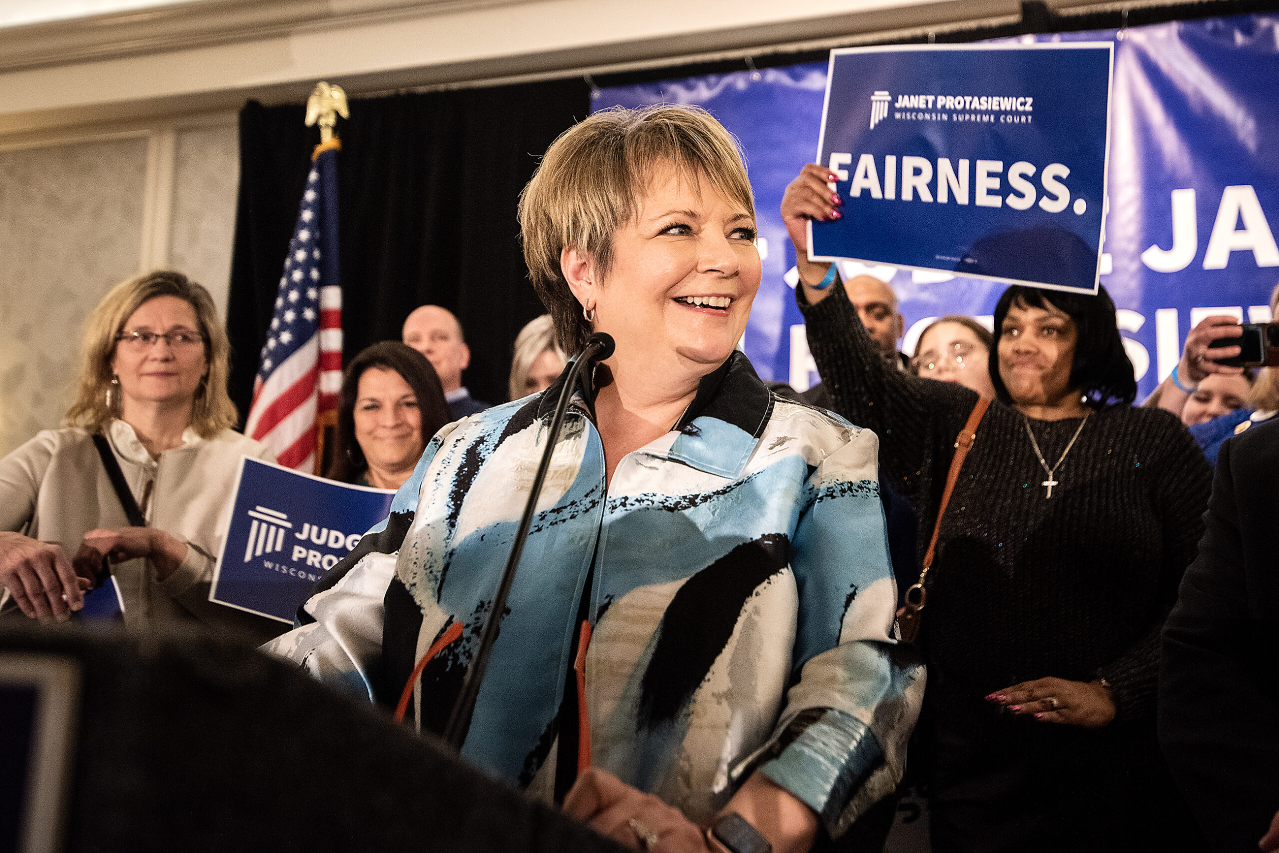 Janet Protasiewicz smiles and looks off at the crowd. A woman in the crowd behind her holds up a sign that says "FAIRNESS."