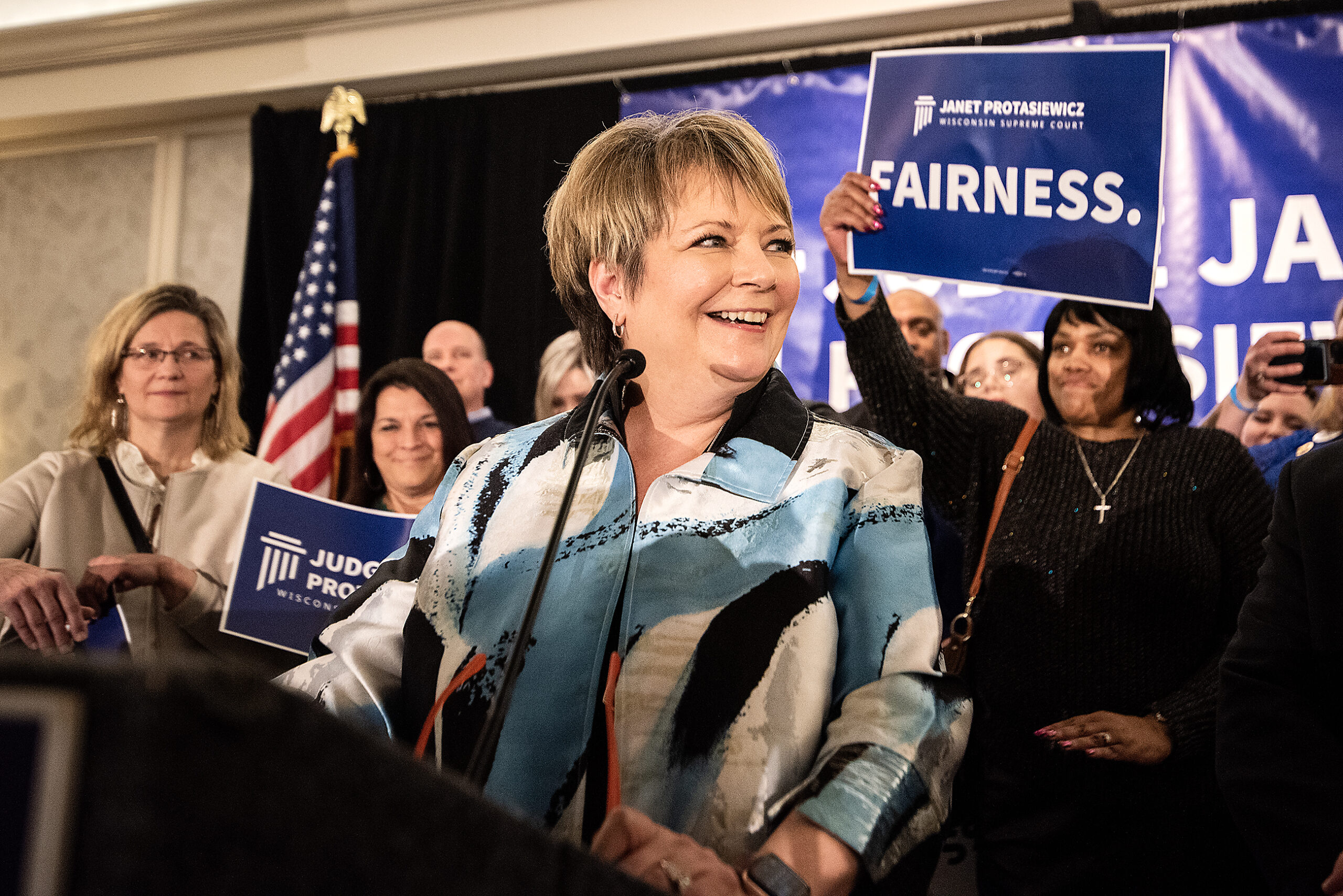 Janet Protasiewicz smiles and looks off at the crowd. A woman in the crowd behind her holds up a sign that says 