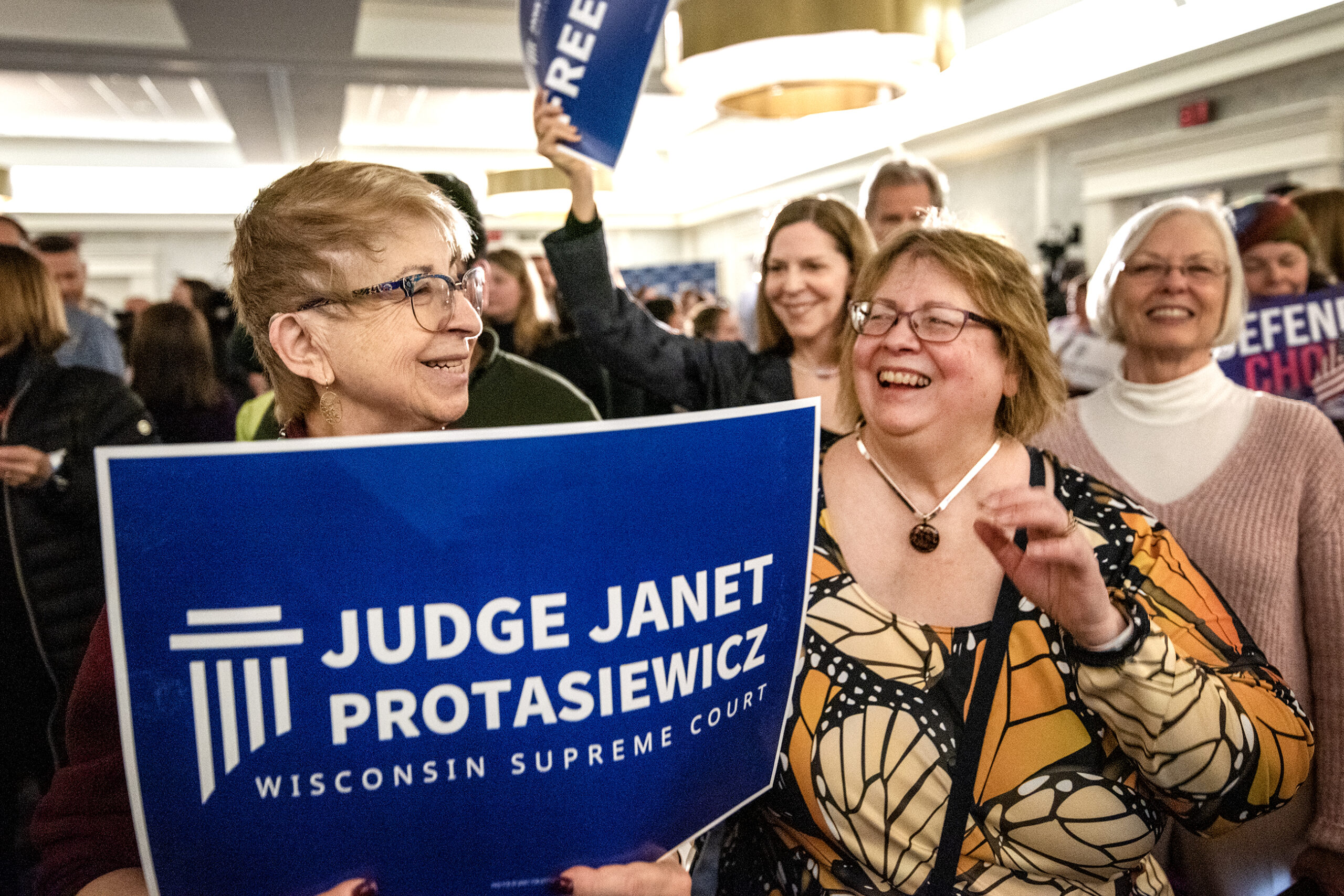 Two women stands together. One holds a blue sign for Judge Janet Protasiewicz