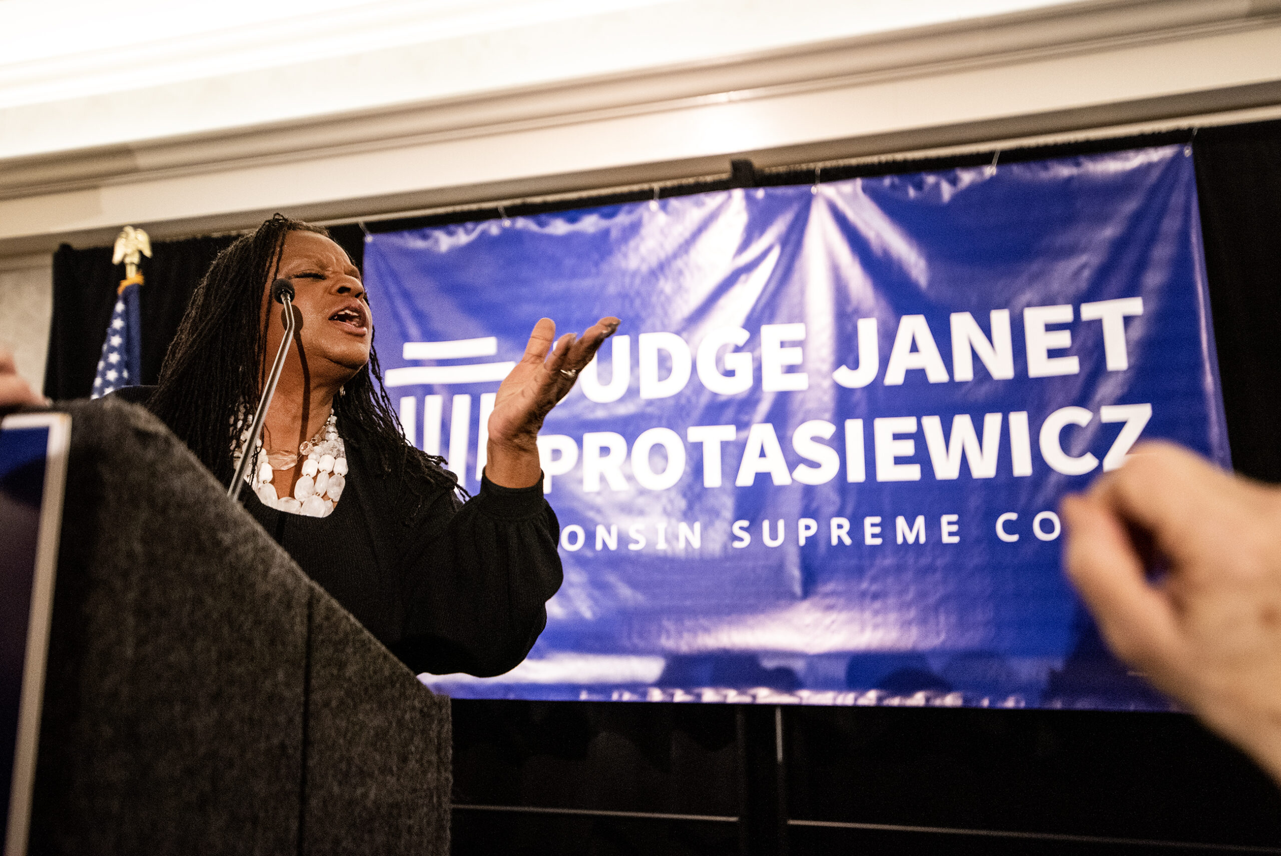 U.S. Rep. Gwen Moore blows a kiss from the podium. A banner for Janet Protasiewicz is seen behind her.