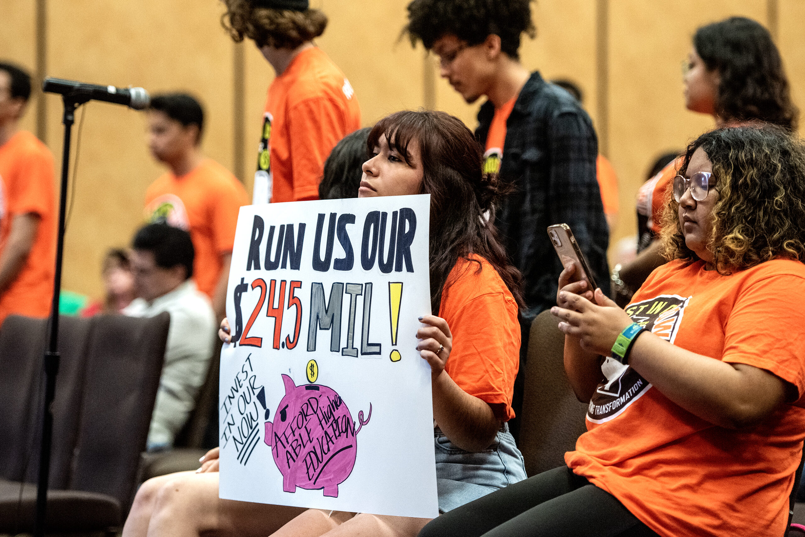 A woman holds a sign that says "Run us our $24.5 mil!"