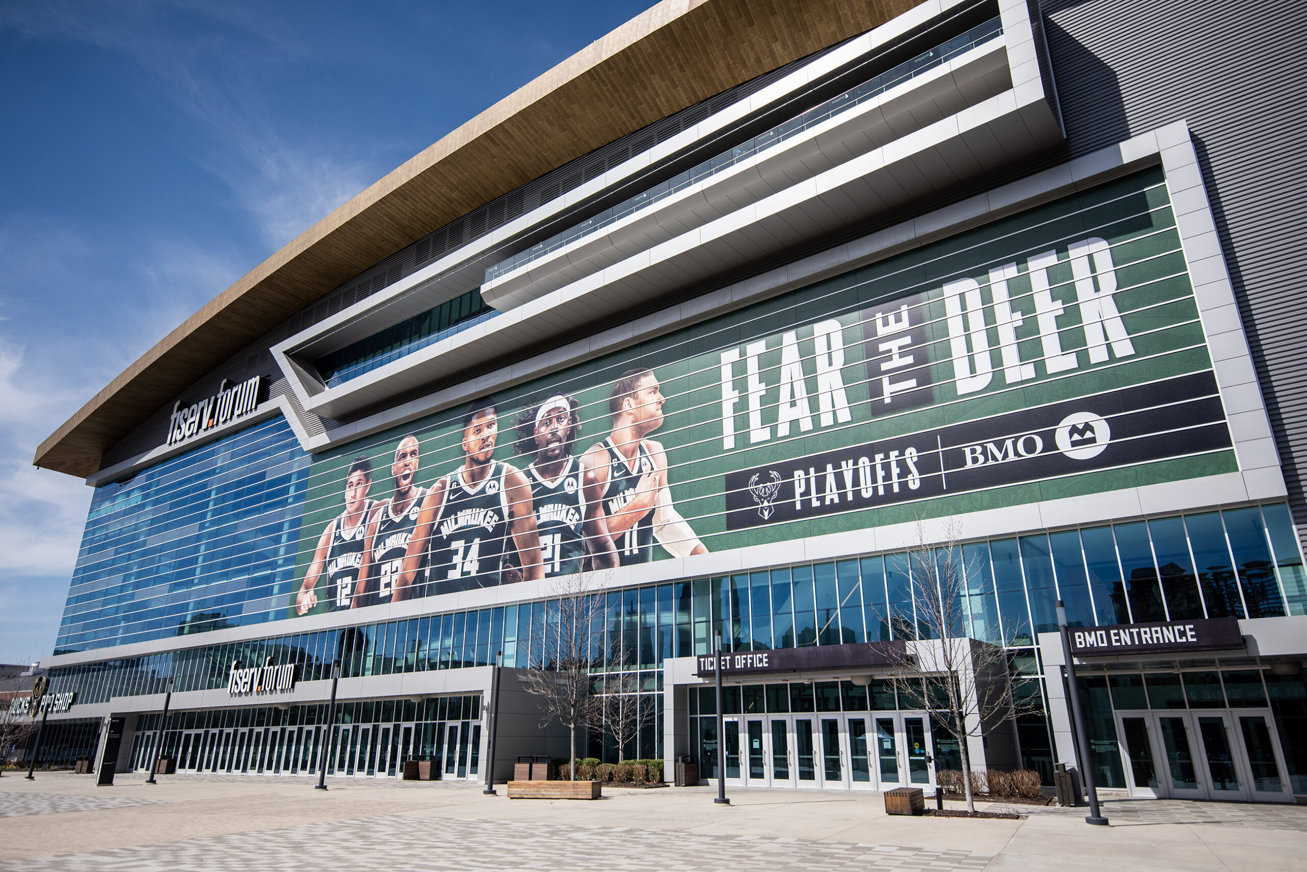 A blue sky is seen behind the curved roof of the Fiserv Forum. Images of 5 players next to the phrase 